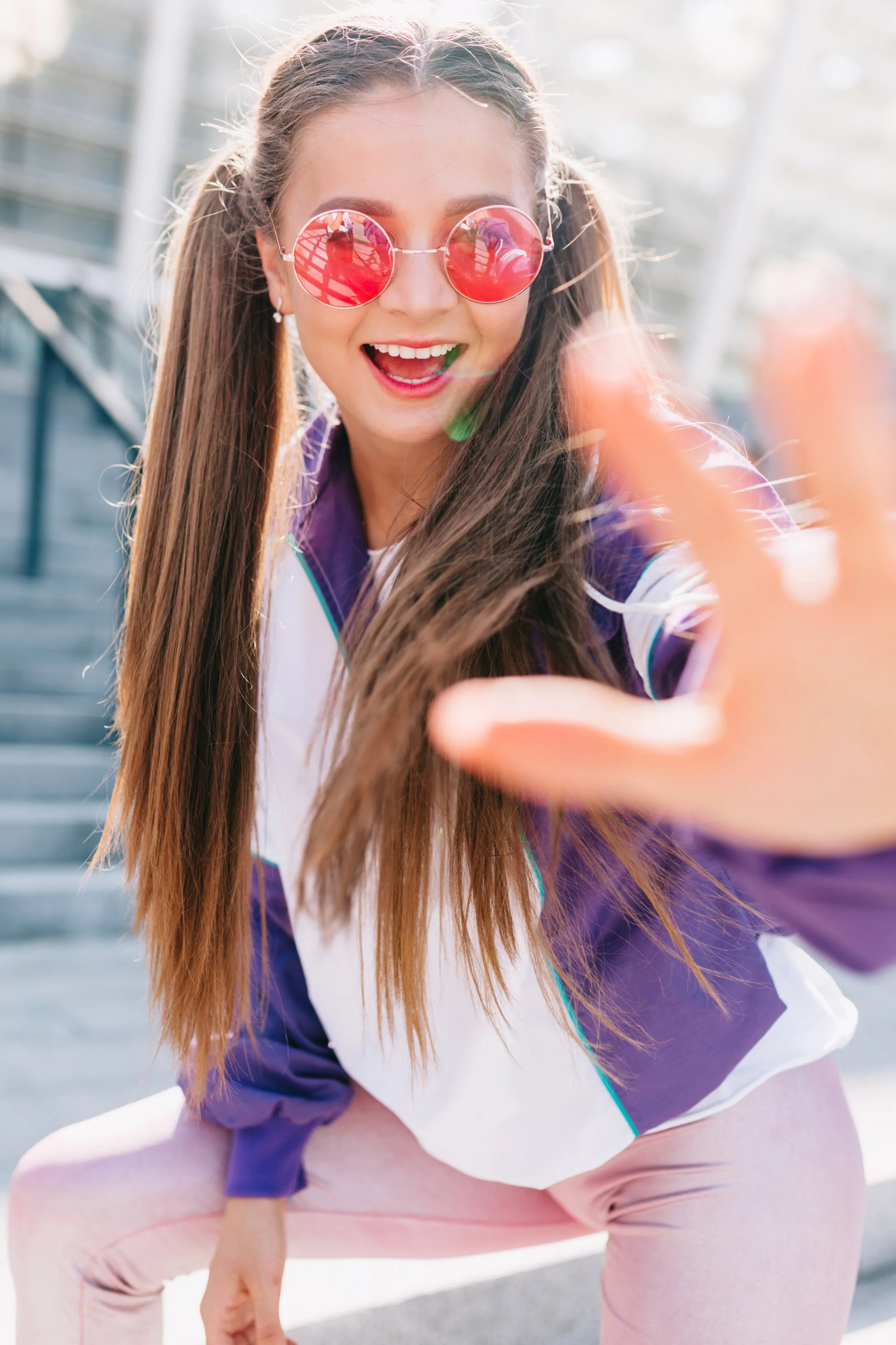 Smiling young woman with long brown hair and pink round sunglasses reaching toward the camera outdoors on stairs.