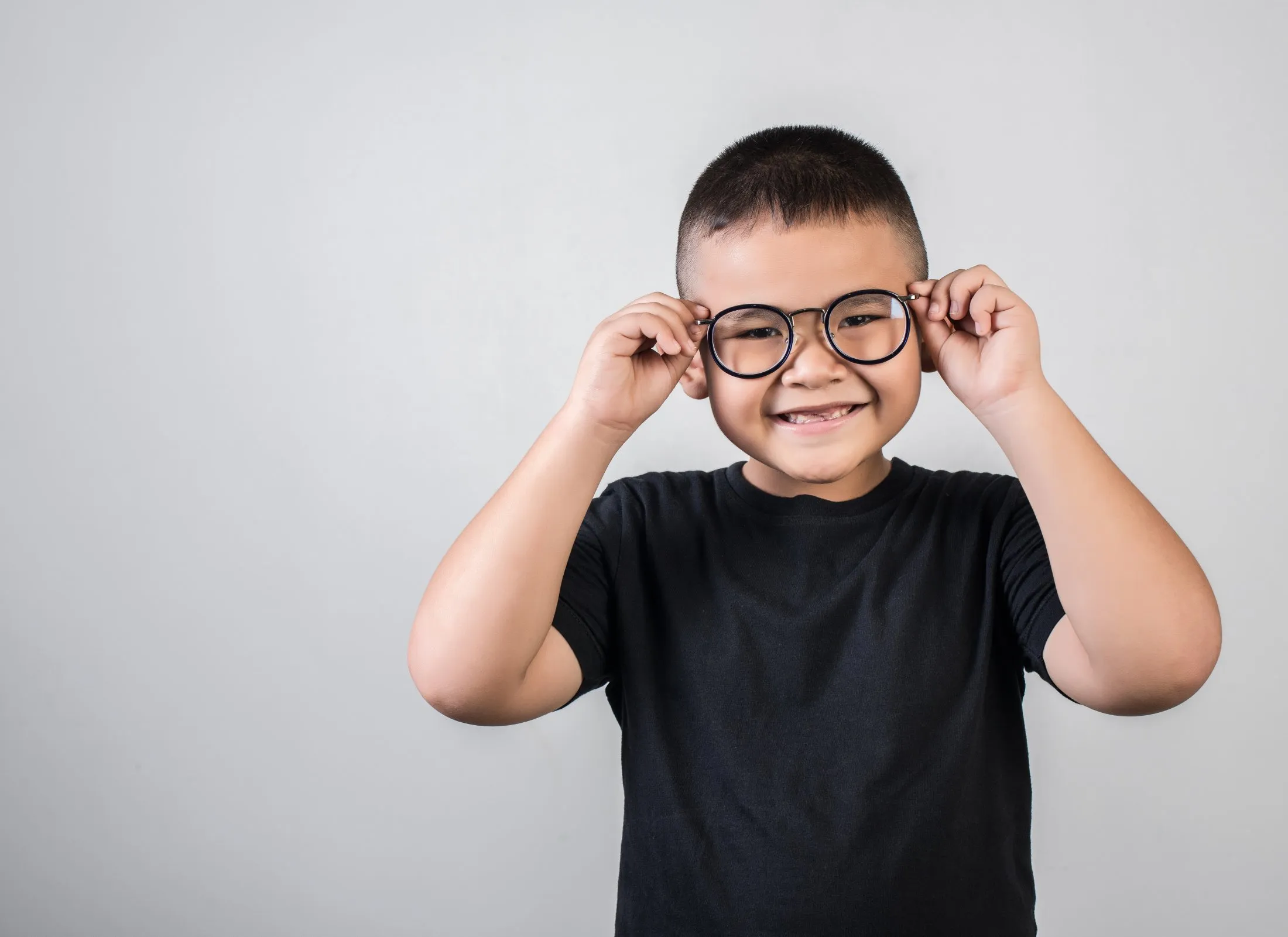 Smiling young boy adjusting round black glasses against a plain gray background.