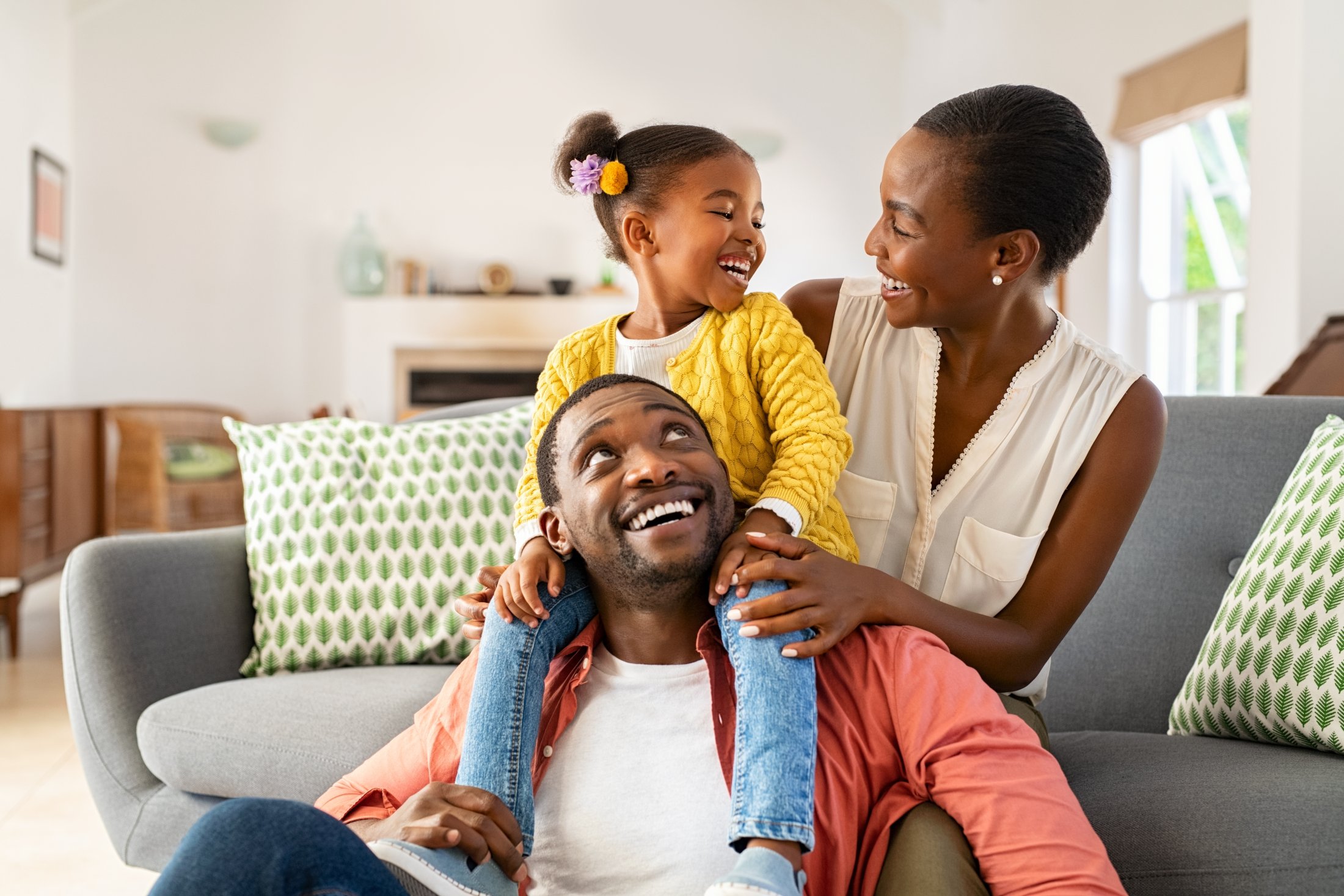 Smiling family with father sitting on the floor holding daughter on his shoulders and mother beside them on a gray sofa in a bright living room.
