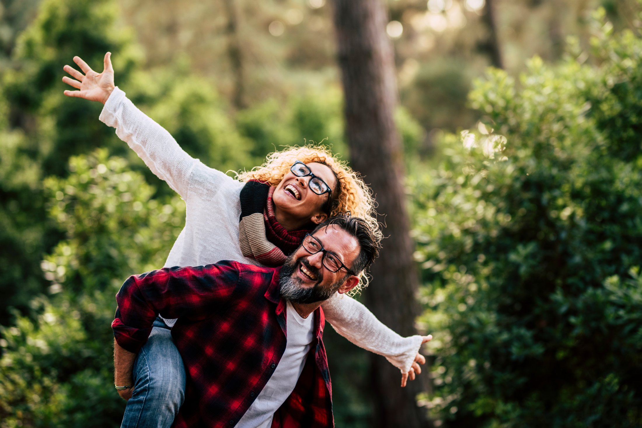 Happy couple outdoors with woman on man's back, both smiling and spreading arms.