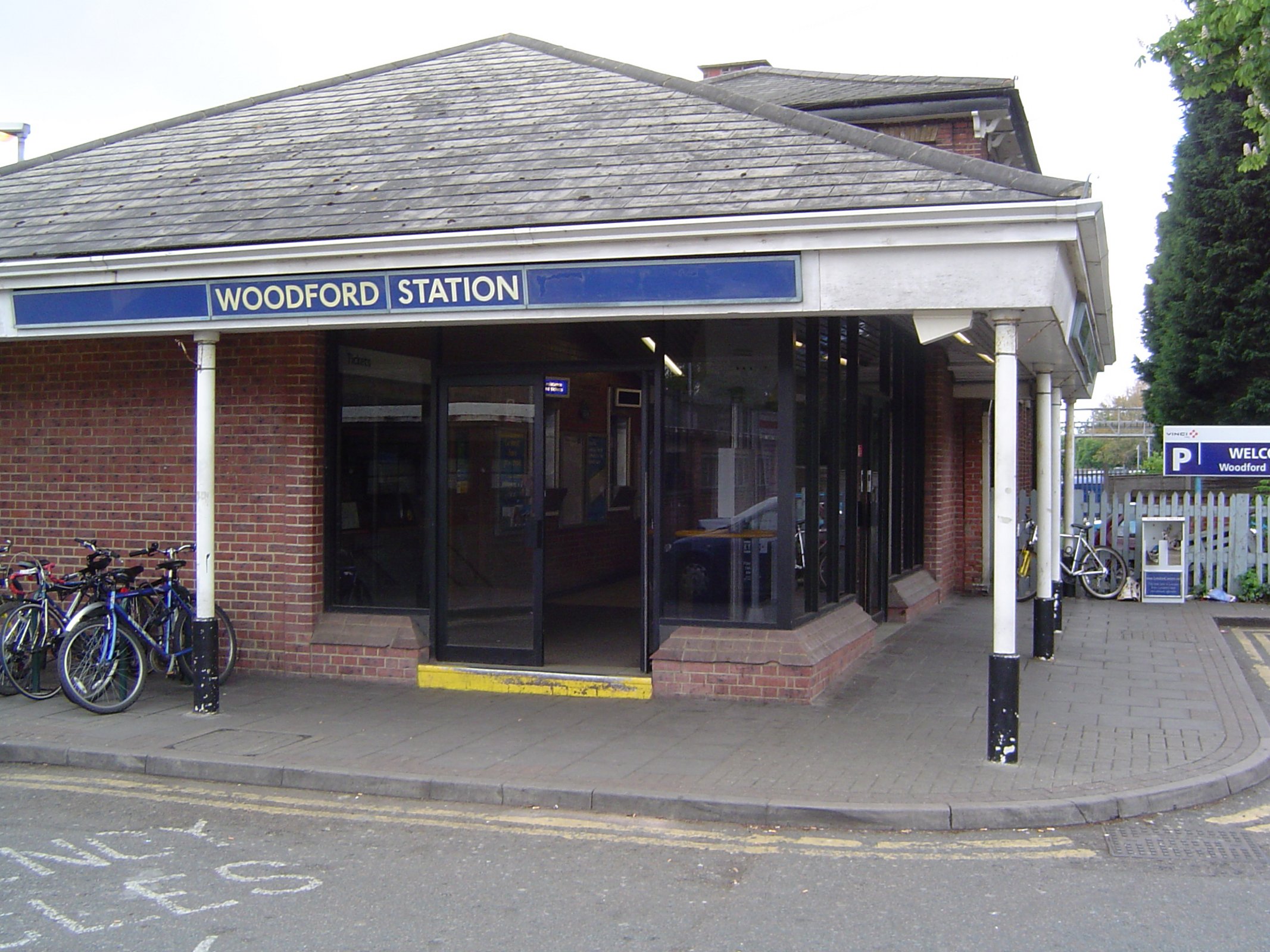 Entrance of Woodford Station with a blue sign above the door and bicycles parked on the left side.