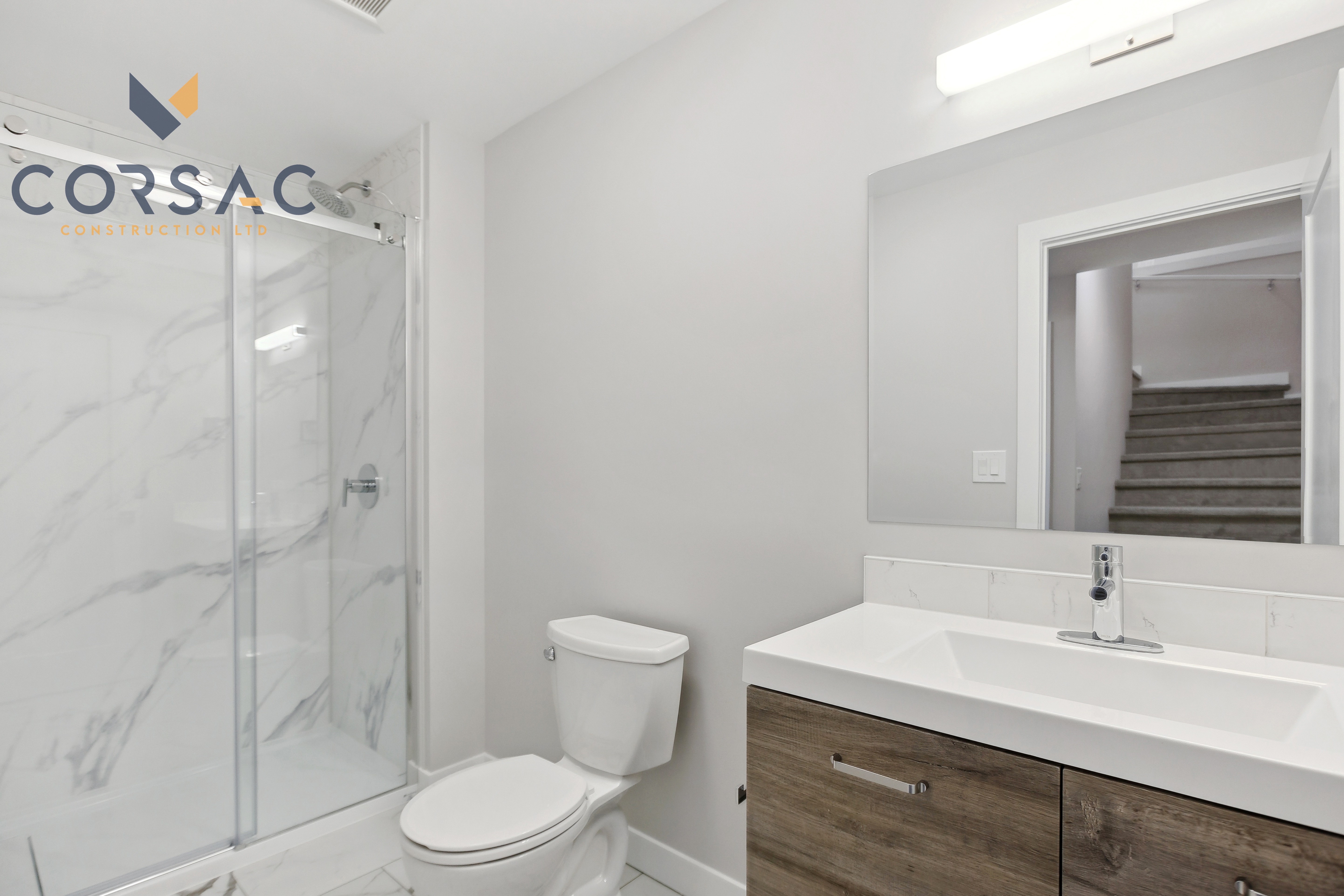 Modern bathroom with glass shower enclosure, white toilet, and wooden vanity with a white sink and large mirror showing stairs in the reflection.