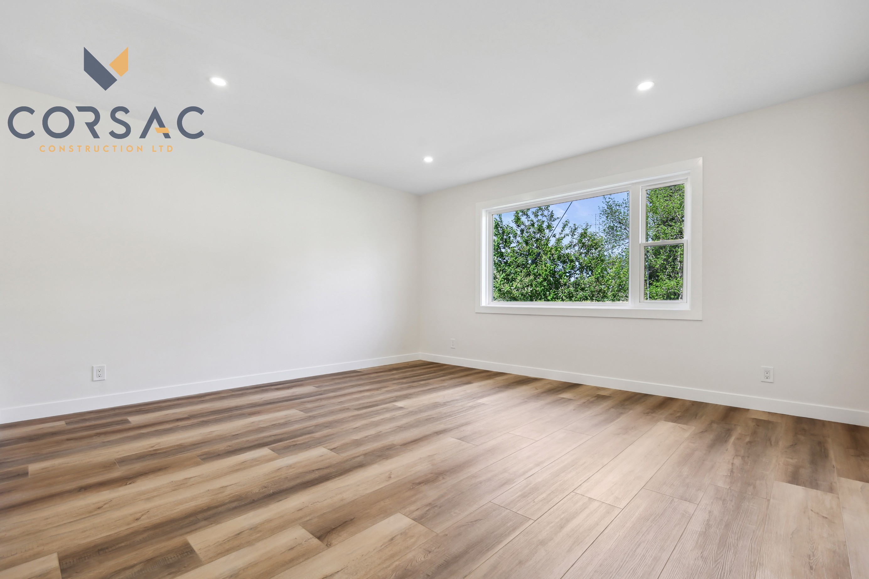 Empty modern room with wood flooring, white walls, recessed lighting, and a large window showing green trees outside.