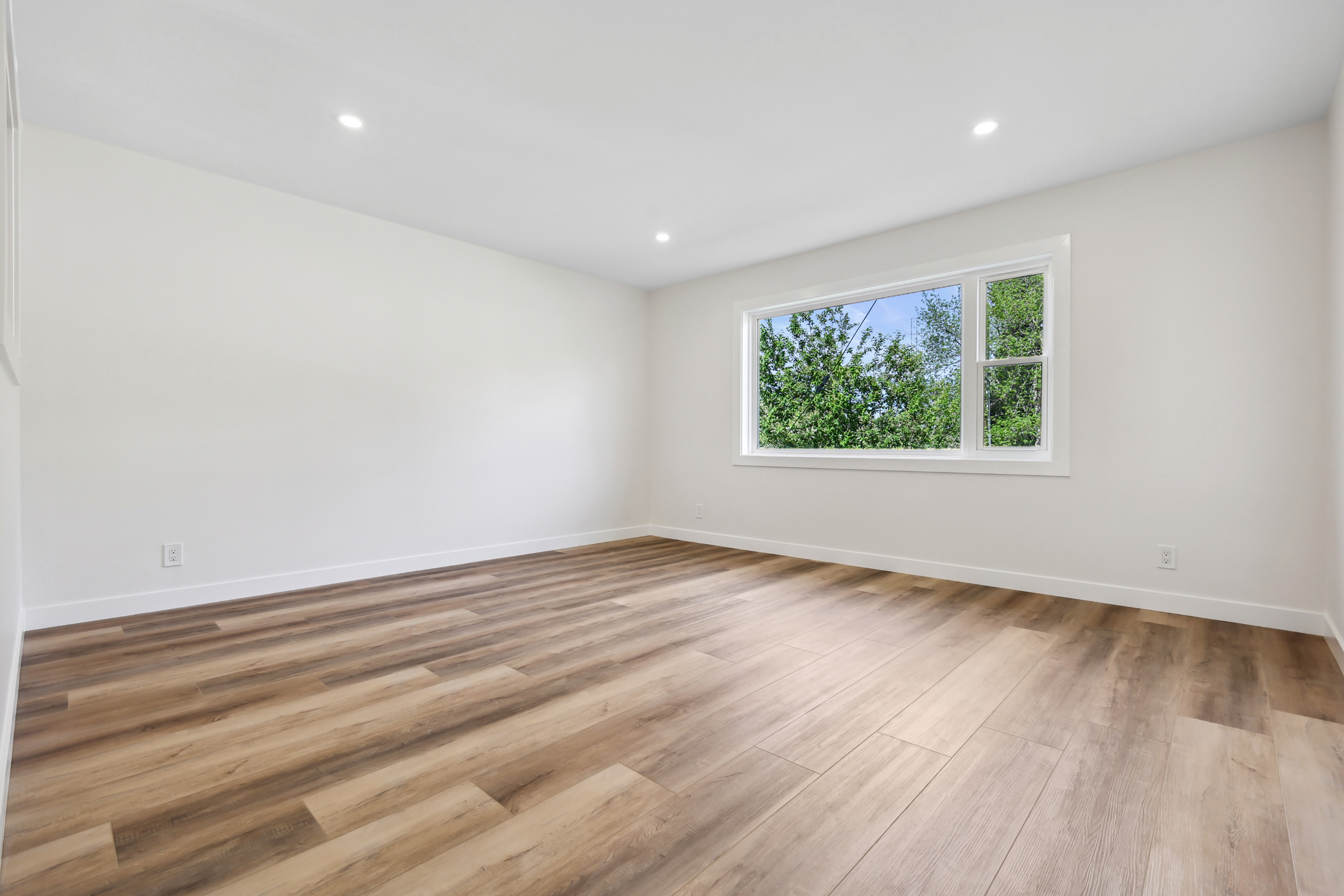 Empty room with light wood flooring, white walls, and a large window showing green trees outside.