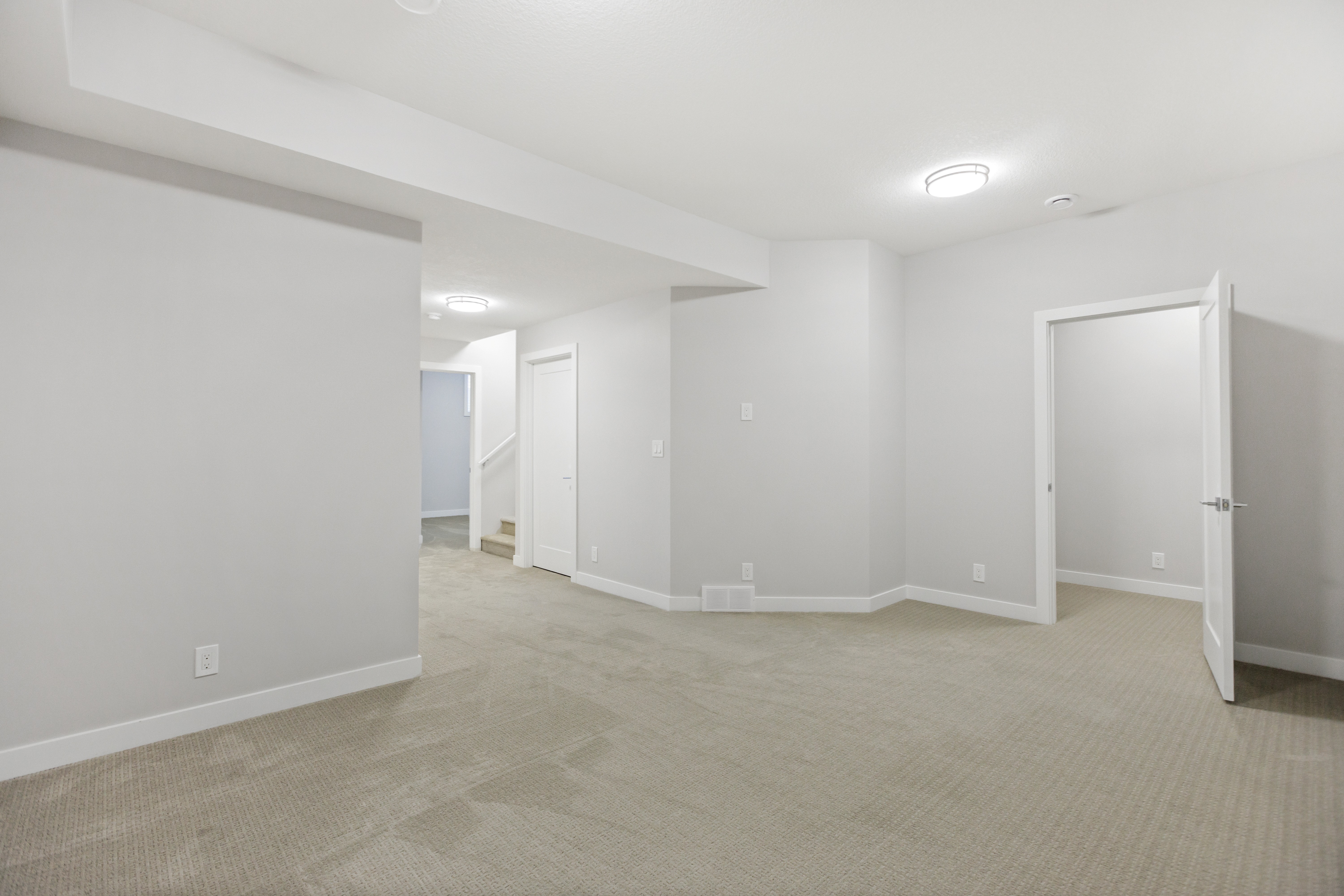 Empty room with beige carpet, white walls, ceiling lights, stairs, and open white door leading to another room.