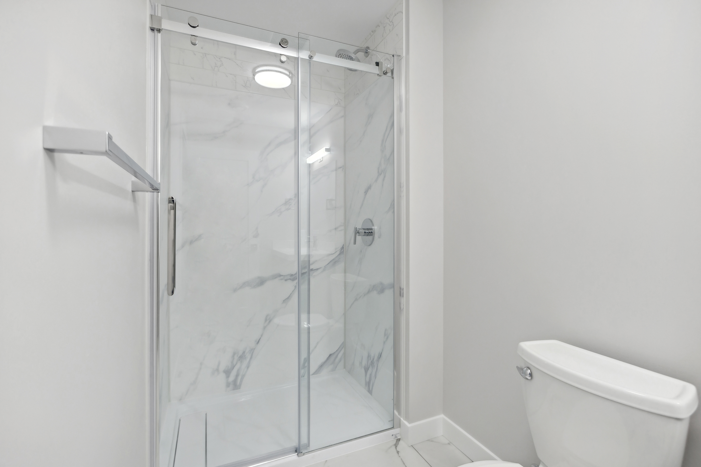 Modern bathroom with a glass sliding door shower featuring marble walls and a white toilet beside it.
