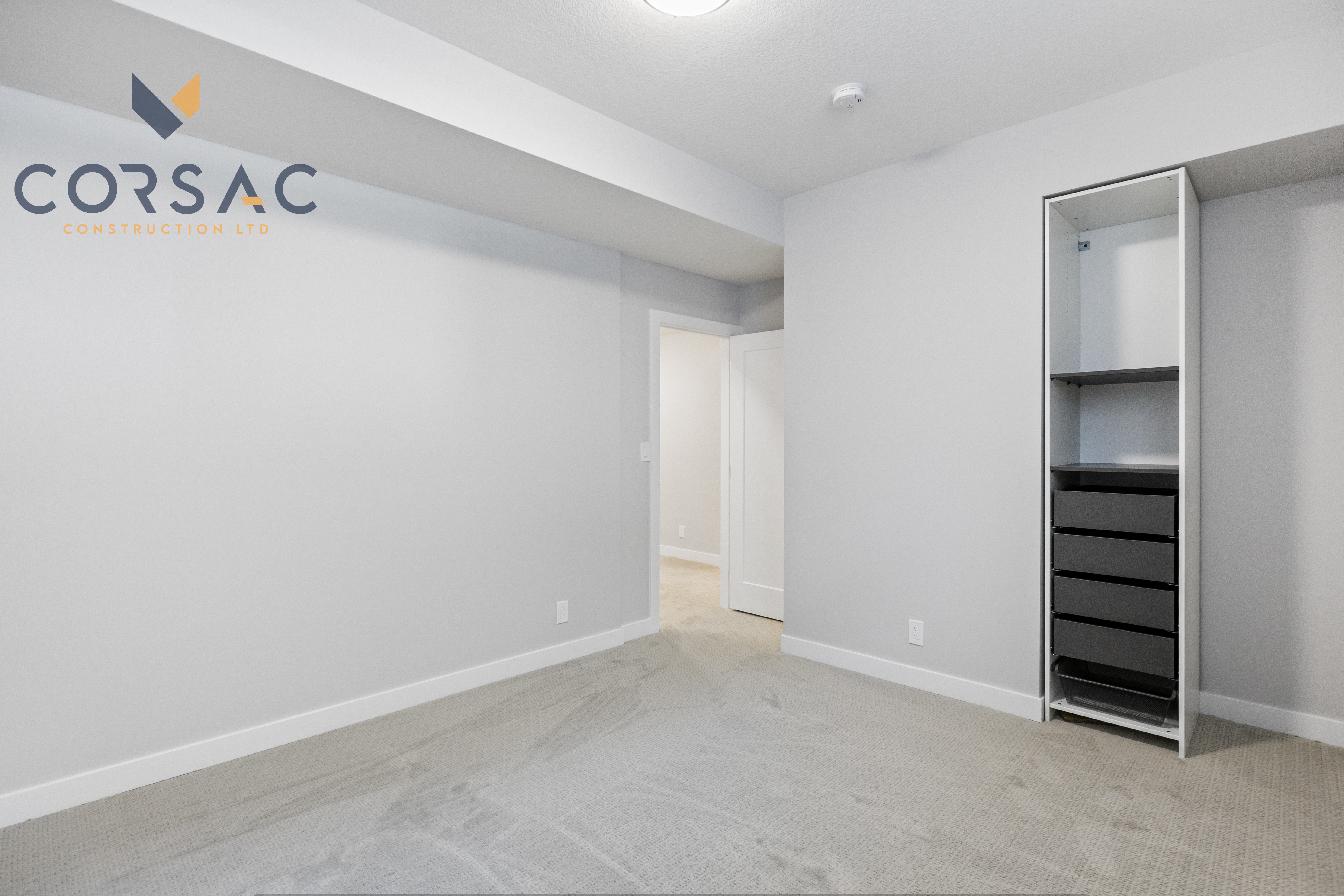 Empty room with light gray walls, beige carpet, an open white door, and a built-in shelving unit with black drawers.