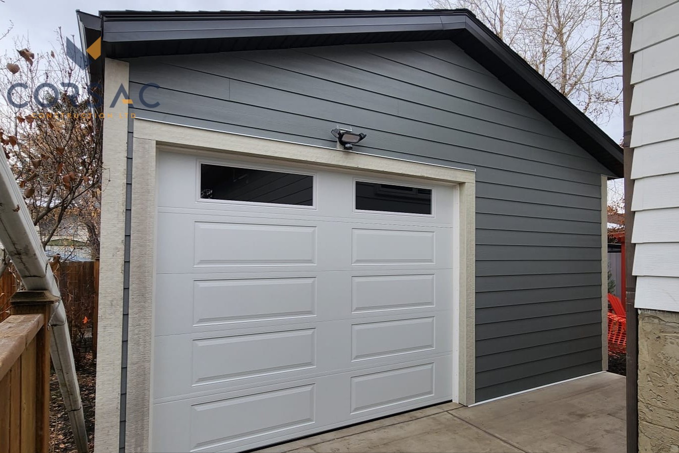 Gray detached garage with white panel garage door and two small windows at the top under an outdoor light.