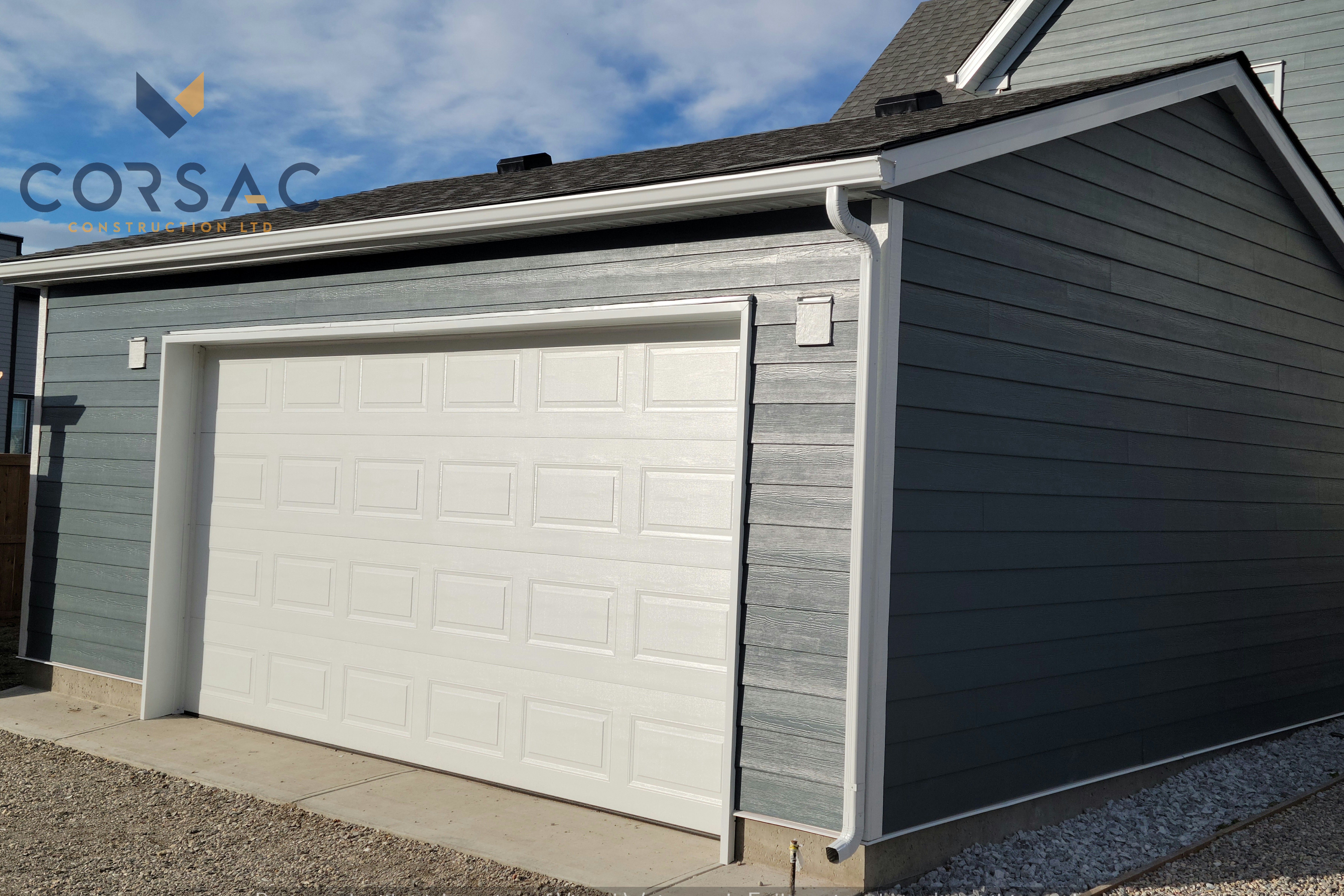 Modern detached garage with white paneled door and gray horizontal siding under a sunny sky.