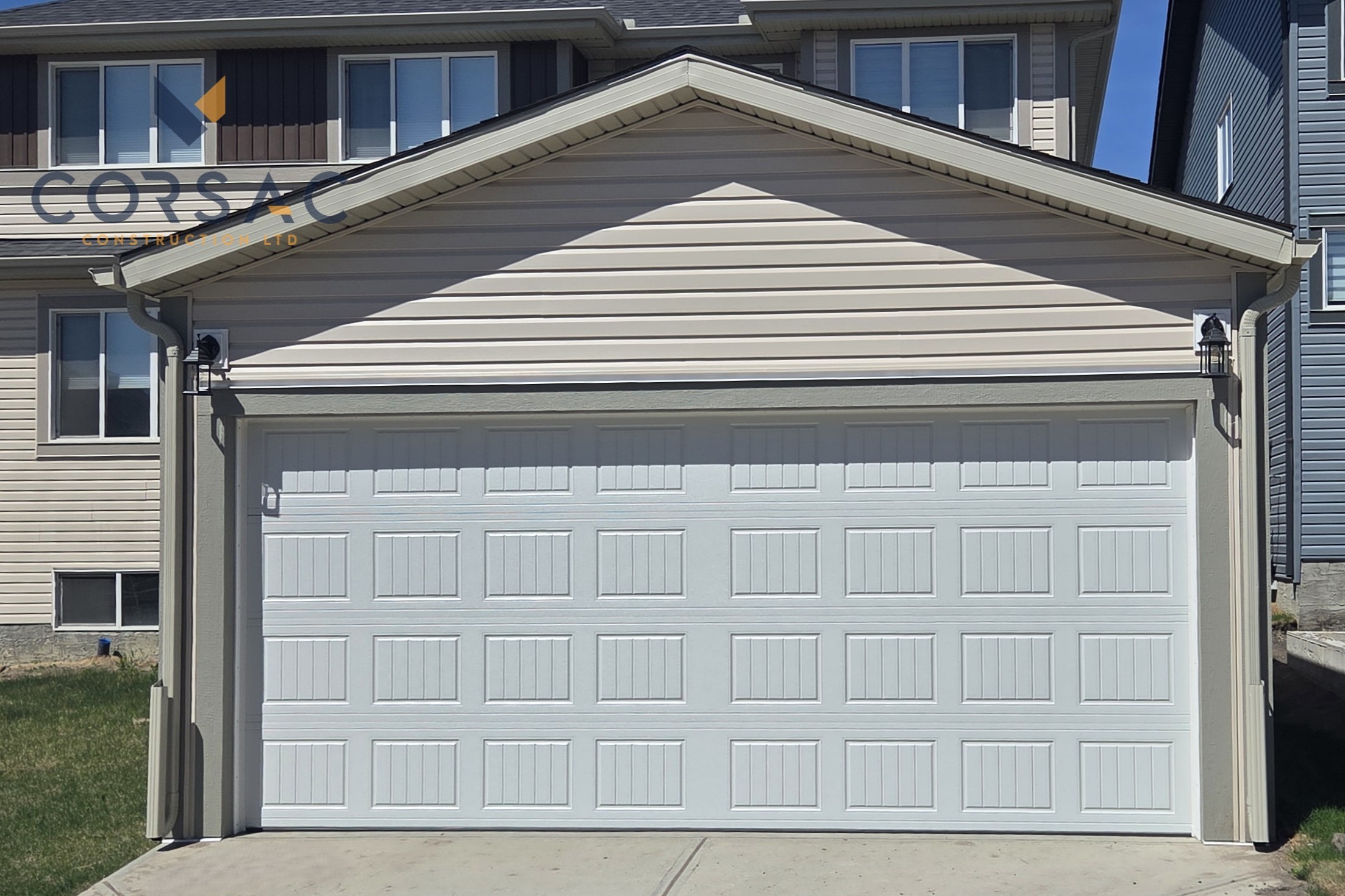Cream-colored detached garage with a white sectional garage door, brown gutters, outdoor light, and sign on the right side under a blue sky.