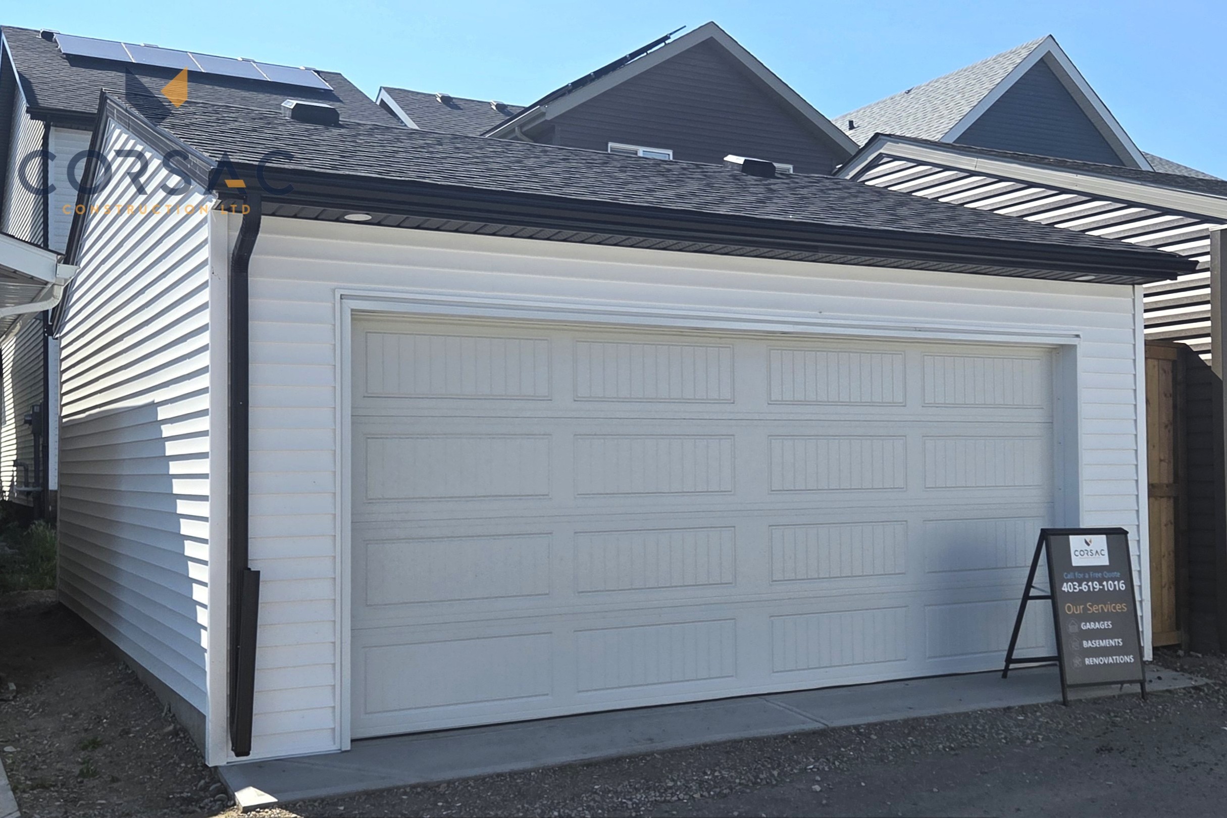 Cream-colored detached garage with a white sectional garage door, brown gutters, outdoor light, and sign on the right side under a blue sky.