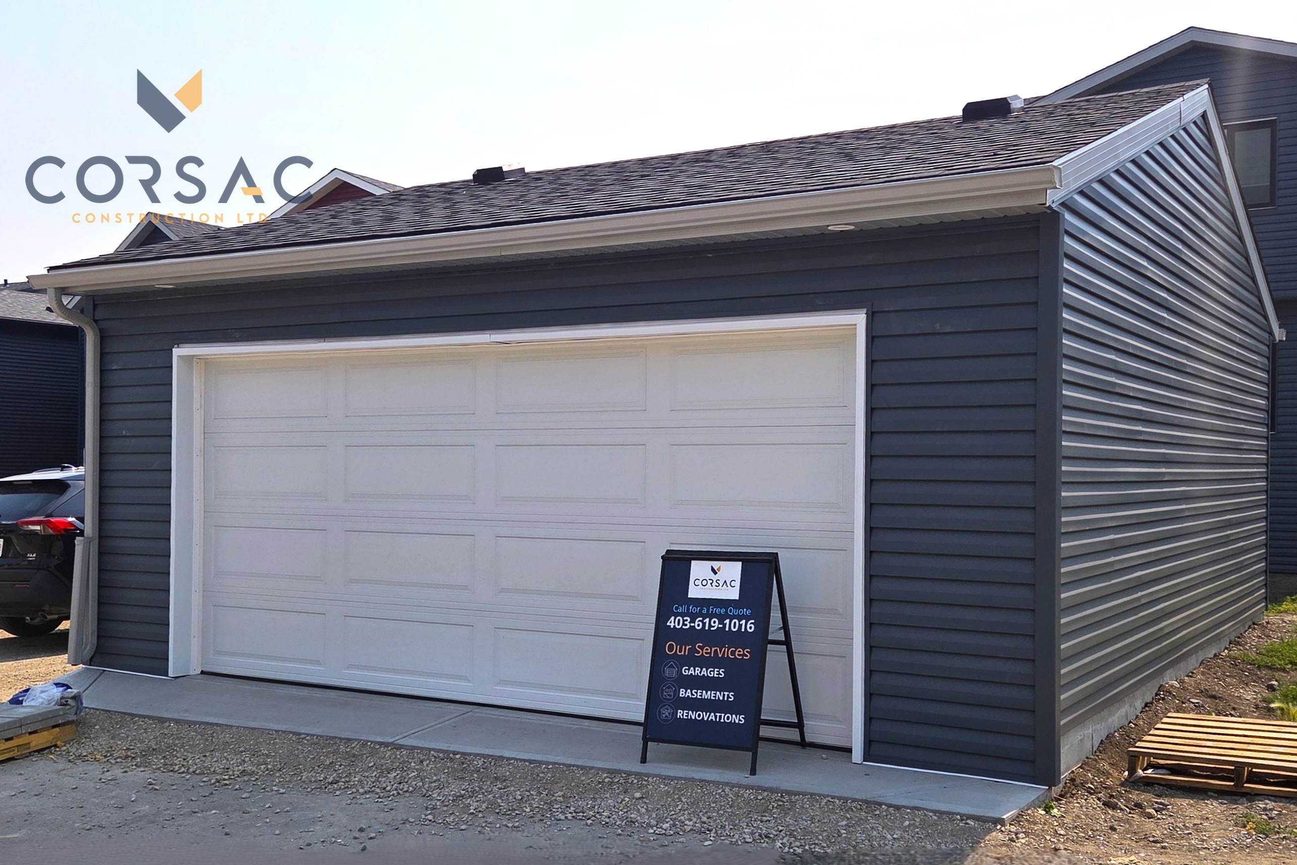Cream-colored detached garage with a white sectional garage door, brown gutters, outdoor light, and sign on the right side under a blue sky.