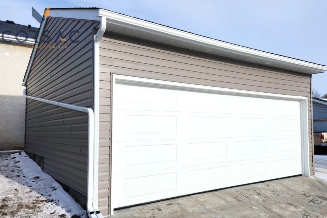 Cream-colored detached garage with a white sectional garage door, brown gutters, outdoor light, and sign on the right side under a blue sky.