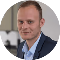 Ein lächelnder Mann in Anzug und Hemd vor unscharfem Hintergrund.