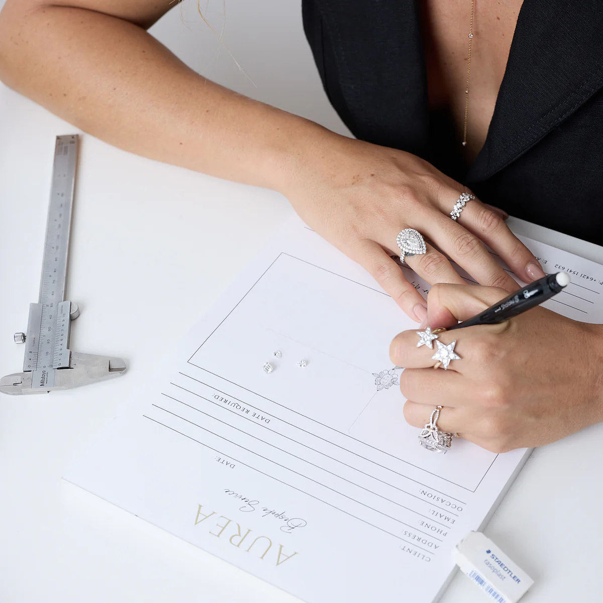 Person wearing multiple diamond rings sketching a jewelry design on paper with a black pen, next to a caliper and an eraser.