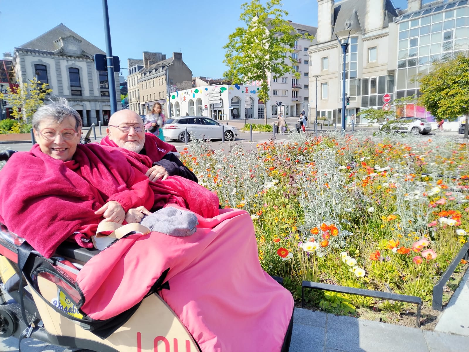 Deux personnes âgées souriantes, enveloppées dans des plaids rouges, installées dans un triporteur adapté devant un parterre de fleurs sauvages, lors d'une sortie en ville.