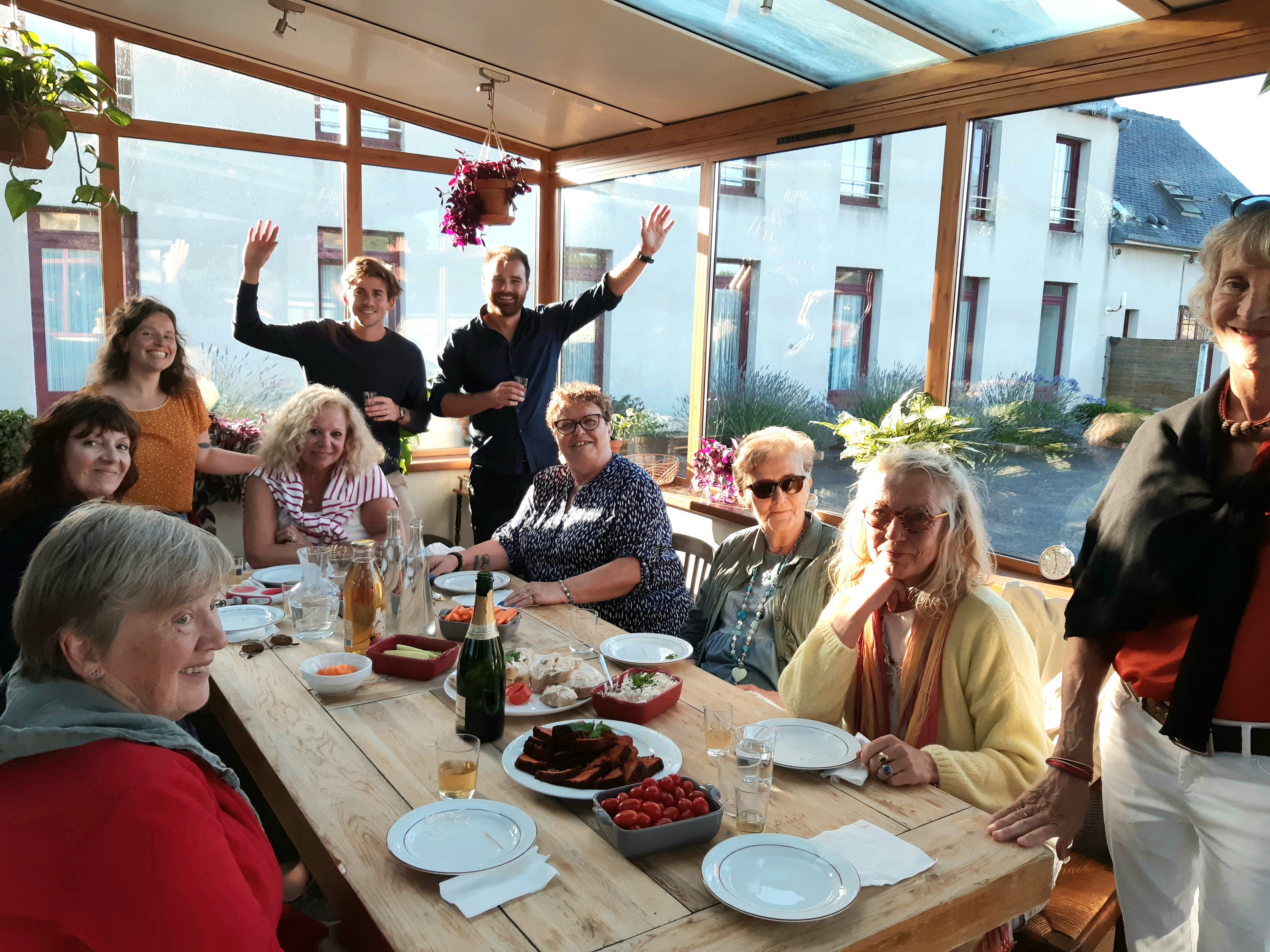 Une dizaine de convives de tous âges partagent un repas convivial autour d'une grande table en bois, dans la véranda lumineuse de l'auberge Les Voisins à Beaucemaine.