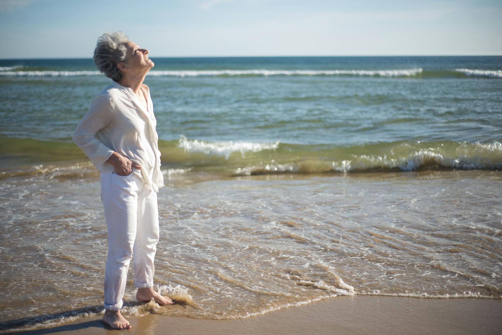 Femme âgée vêtue de blanc, pieds nus au bord de l'eau sur une plage, les yeux fermés et le visage tourné vers le soleil, respirant l'air marin lors d'un moment de ressourcement.
