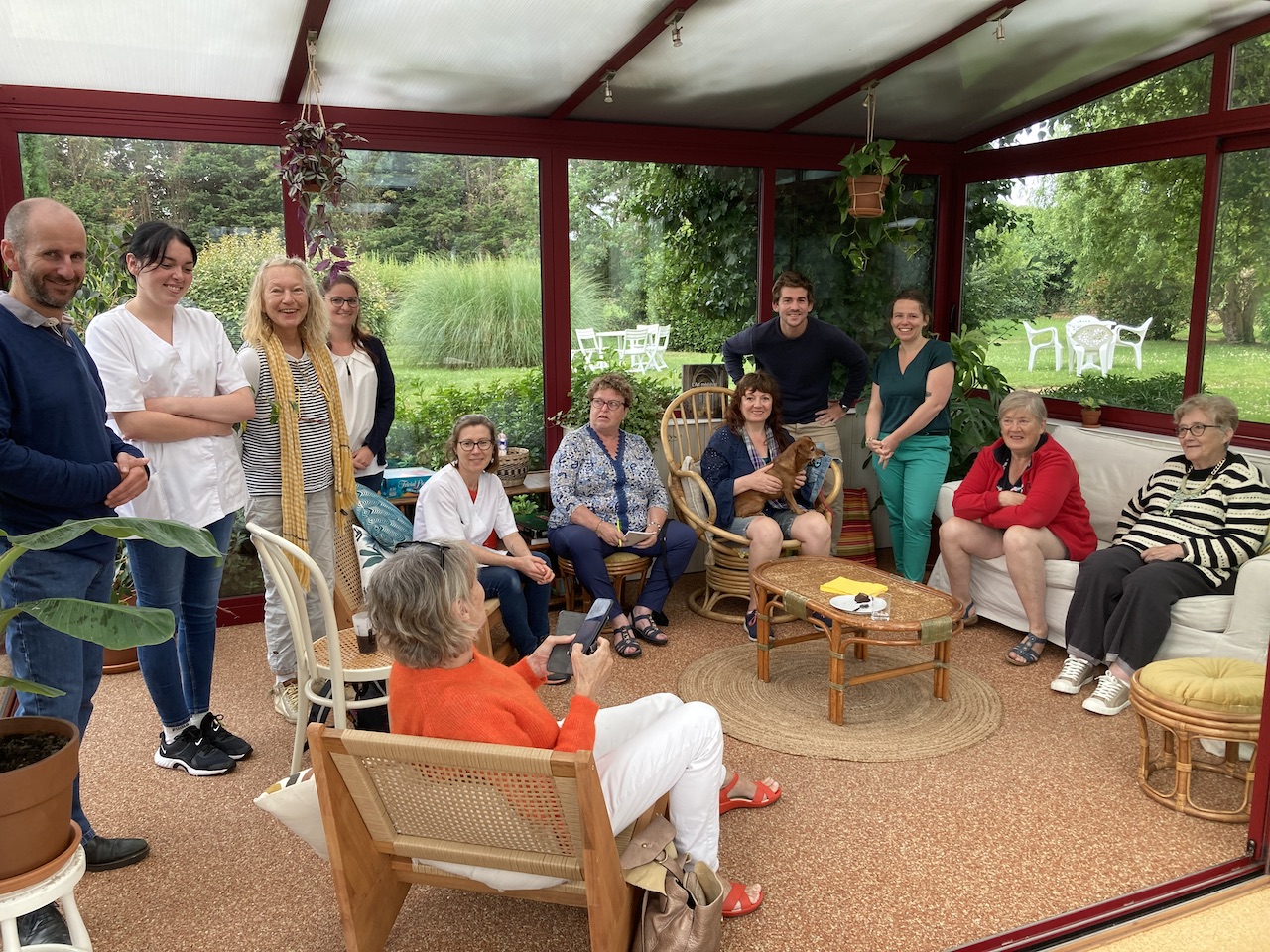 Groupe de résidents, de proches et de membres de l'équipe réunis dans la véranda de l'auberge Les Voisins à Beaucemaine, illustration d'un hébergement inclusif.