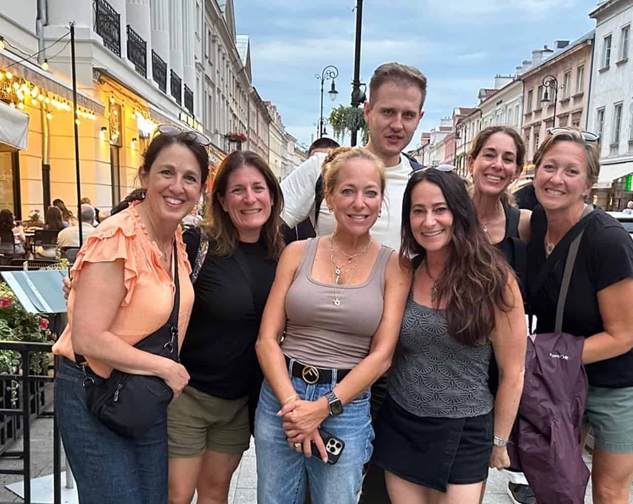 Group of six smiling adults posing together on a city street with historic buildings and outdoor dining in the background.