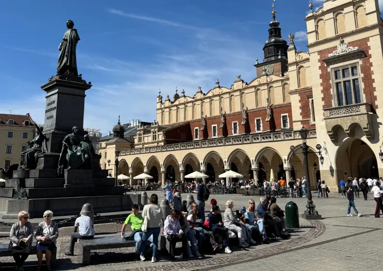 People sitting on benches and walking in a sunny historic square in front of a large building with arches and outdoor cafés.