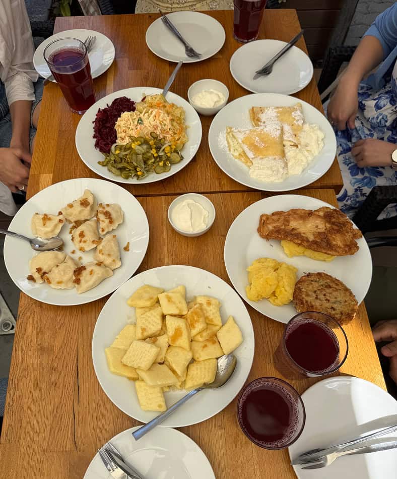 Wooden table with plates of Eastern European dishes including pierogi with onions, potato pancakes, beet salad, pickles, fried cheese curds, and three glasses of red juice.