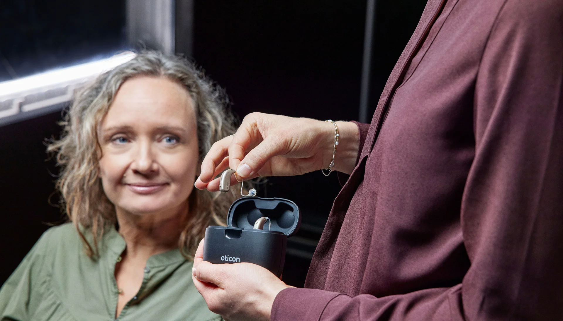 Une personne tenant un appareil auditif Oticon au-dessus de son étui de chargement, une femme aux cheveux bouclés et blonds regardant avec un sourire.