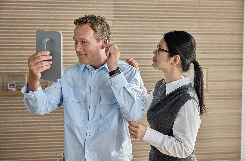 Un homme regarde dans un miroir tout en ajustant un appareil auditif, assisté par une femme portant des lunettes.