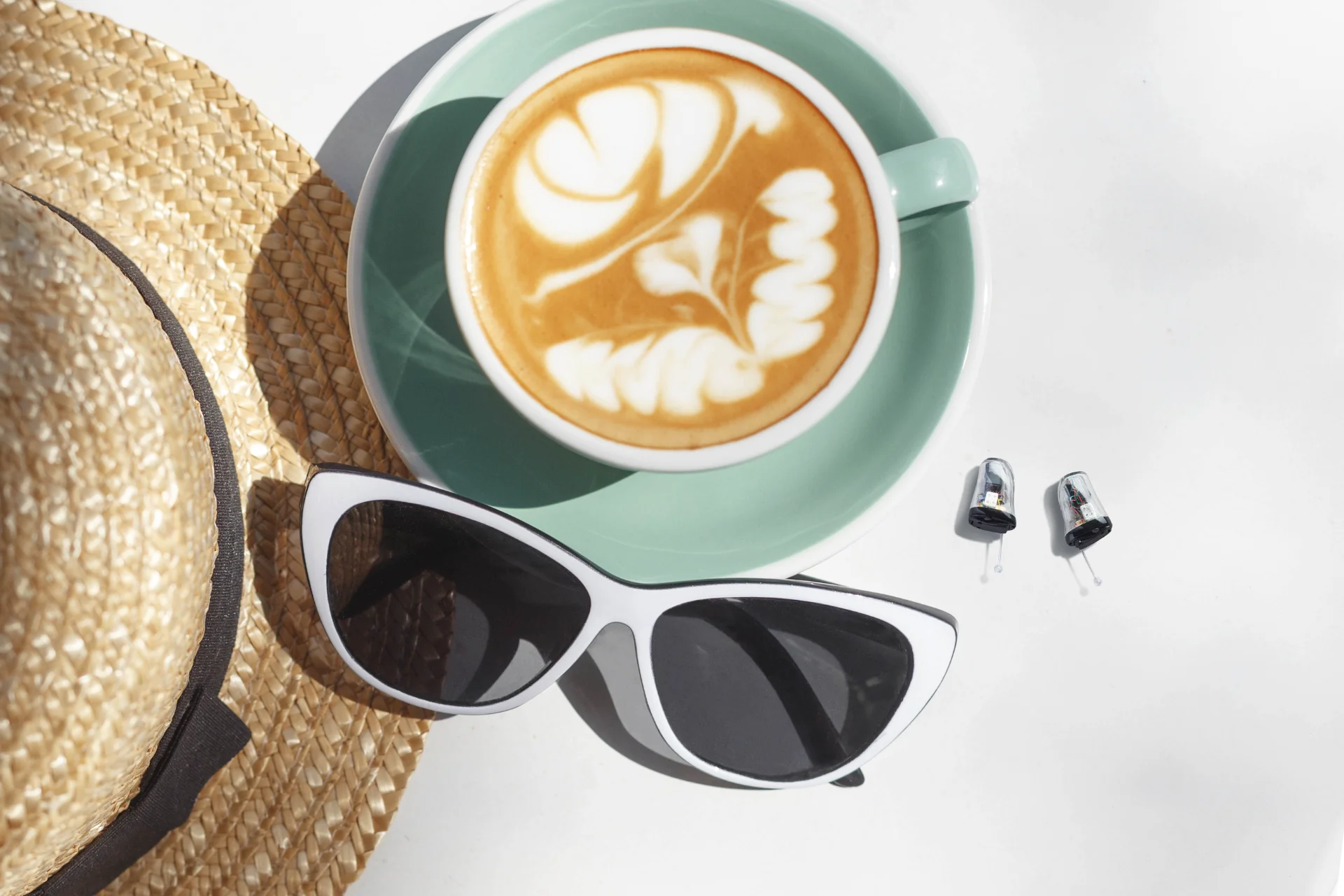 Chapeau de paille, lunettes de soleil blanches, tasse de café latte avec motif de mousse, et deux petits appareils auditifs sur une surface blanche.