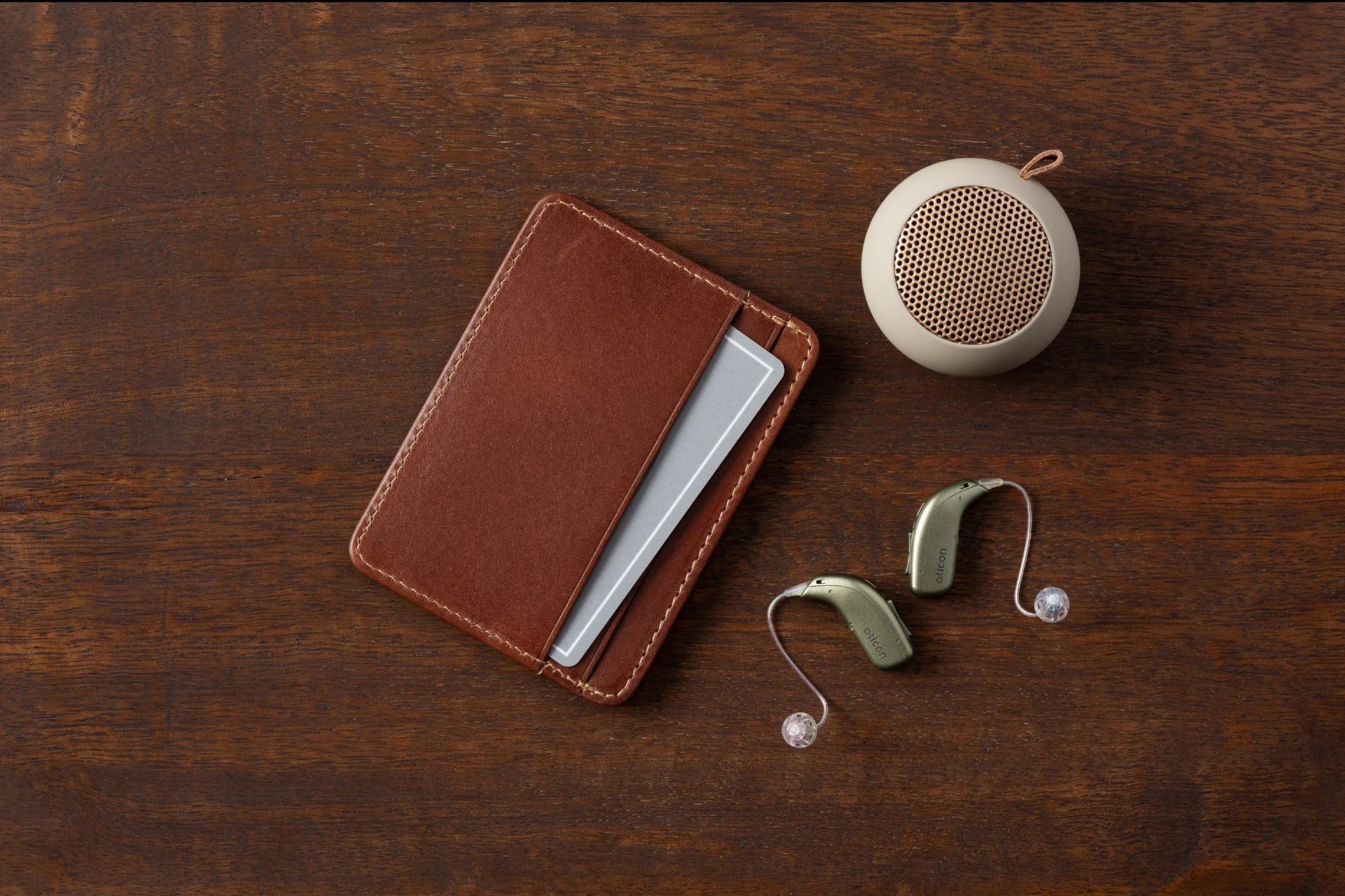 Un portefeuille en cuir marron avec une carte grise partiellement visible, deux aides auditives vertes de marque Oticon et un petit haut-parleur portable rond posé sur une table en bois.