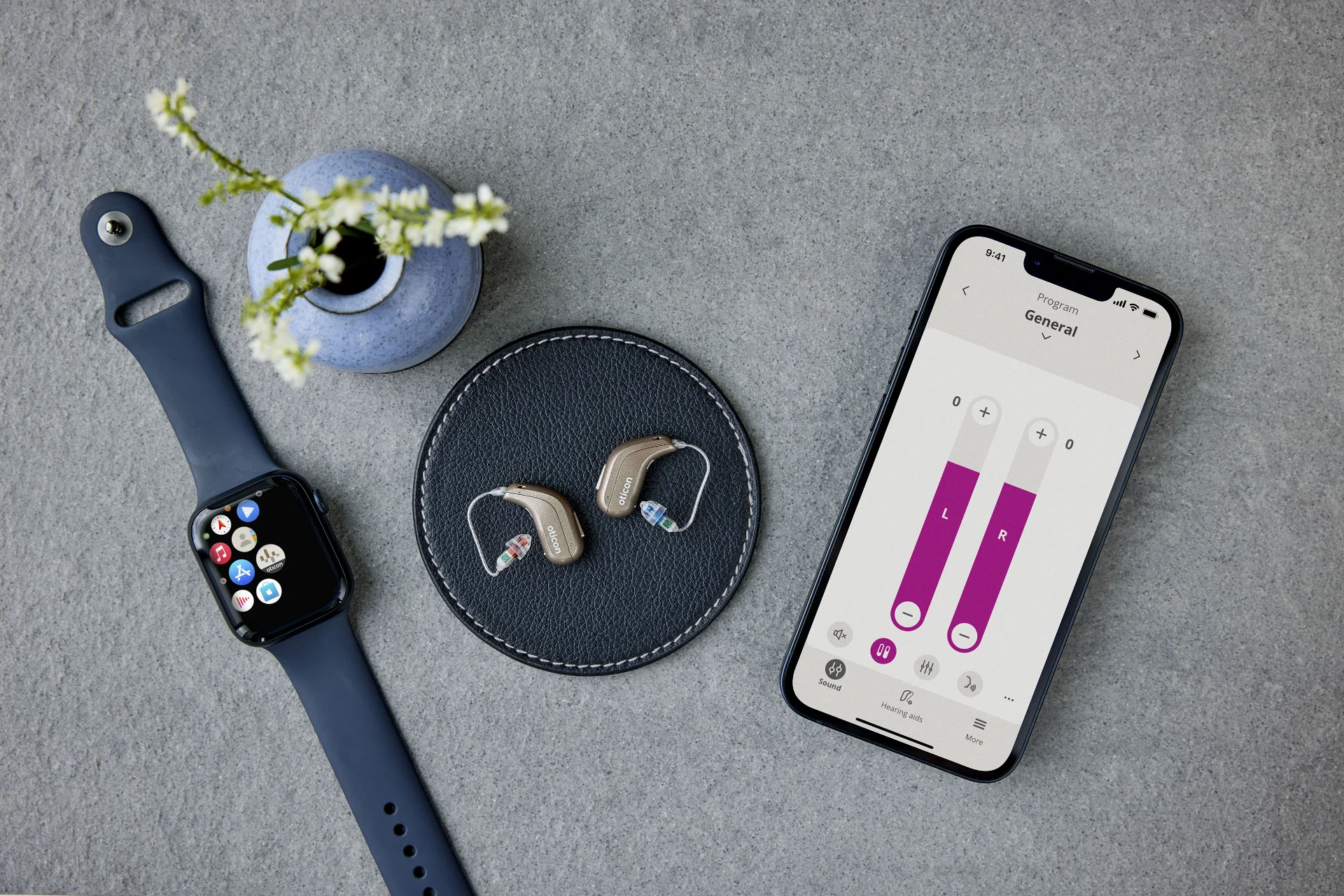 Aides auditives Oticon dans un étui de chargement ouvert posées sur une table en bois avec des lunettes de soleil et un porte-cartes en arrière-plan.