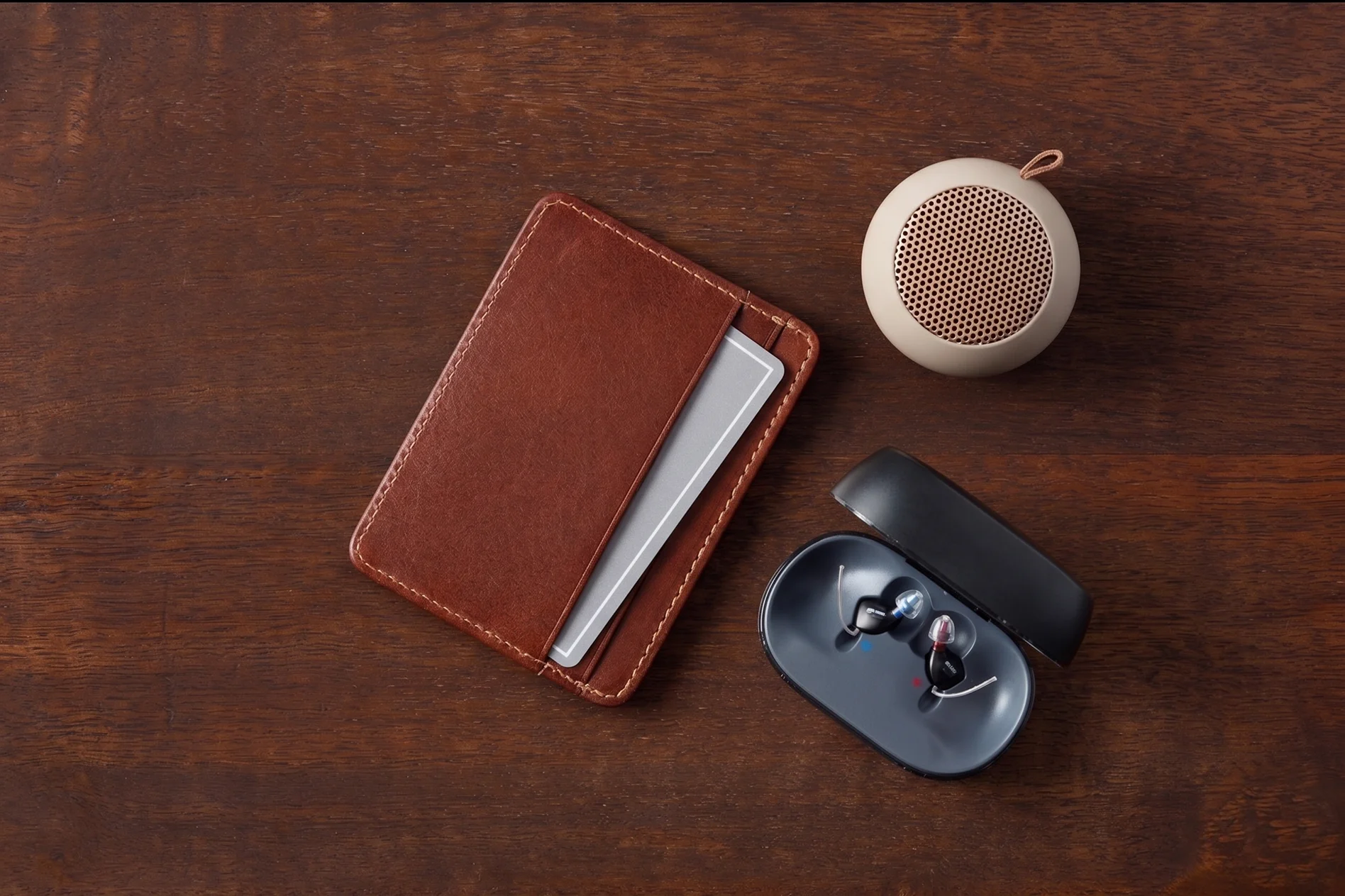 Un portefeuille en cuir marron avec une carte grise partiellement visible, deux aides auditives vertes de marque Oticon et un petit haut-parleur portable rond posé sur une table en bois.