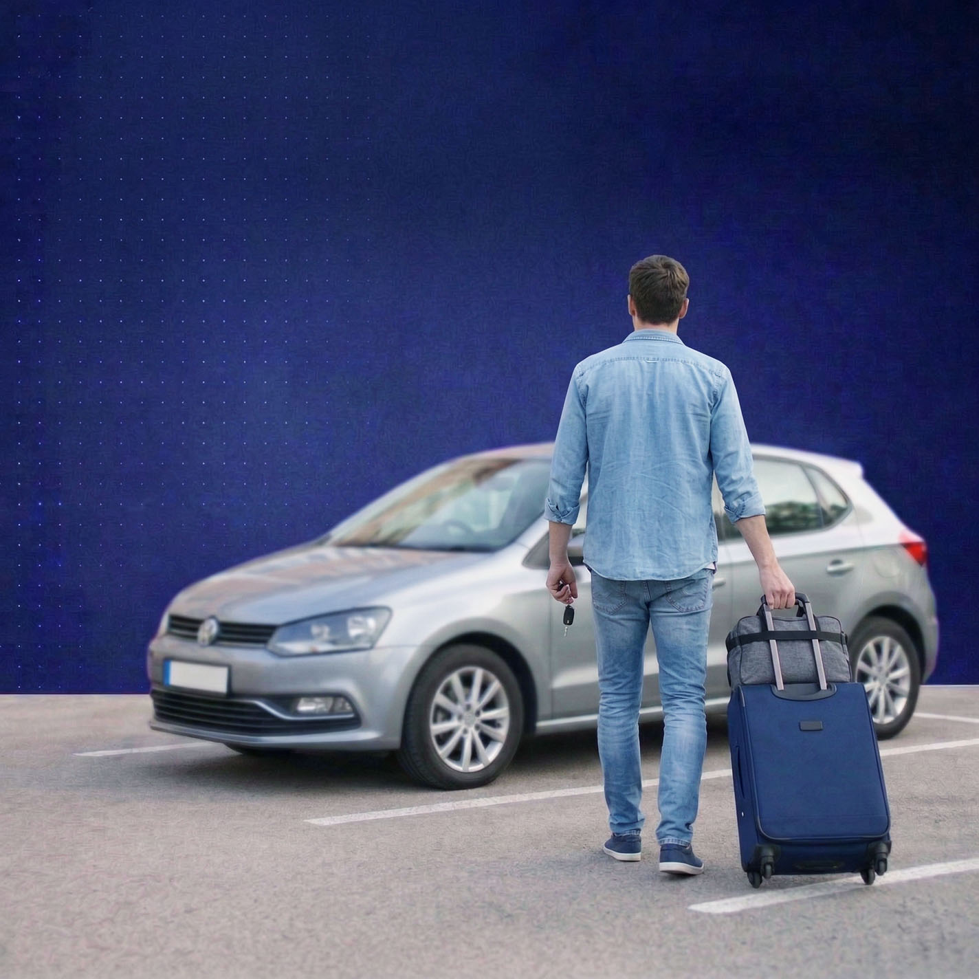 Man in denim outfit holding car keys and pulling a suitcase towards a silver hatchback car in a parking lot.