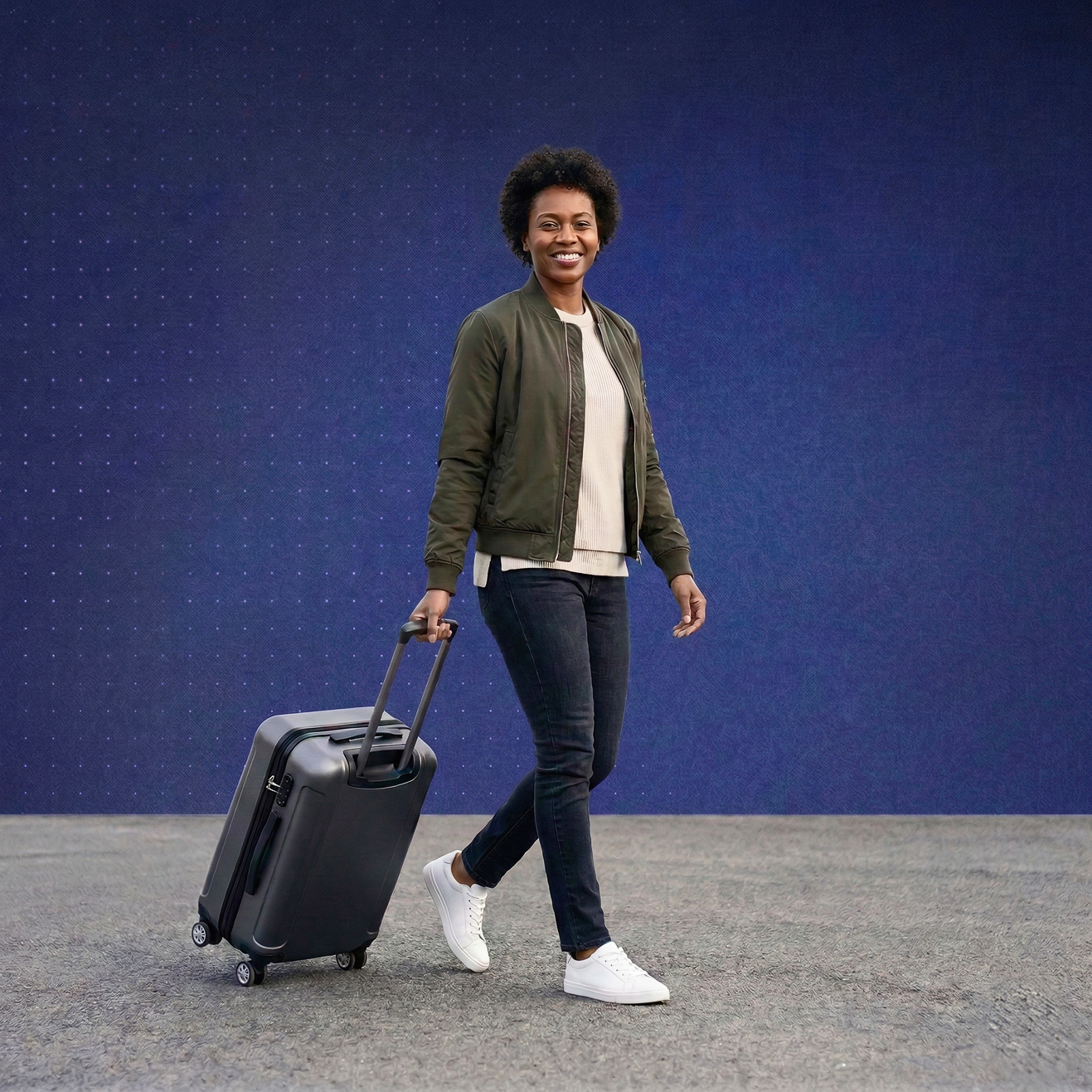 Smiling woman in a jacket walking with a black rolling suitcase on a paved surface against a blue background.