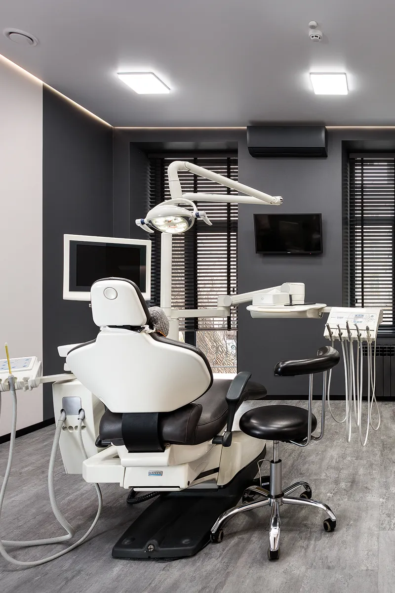 Modern dental treatment room with dental chair, overhead light, and equipment against dark gray walls and wood floor.