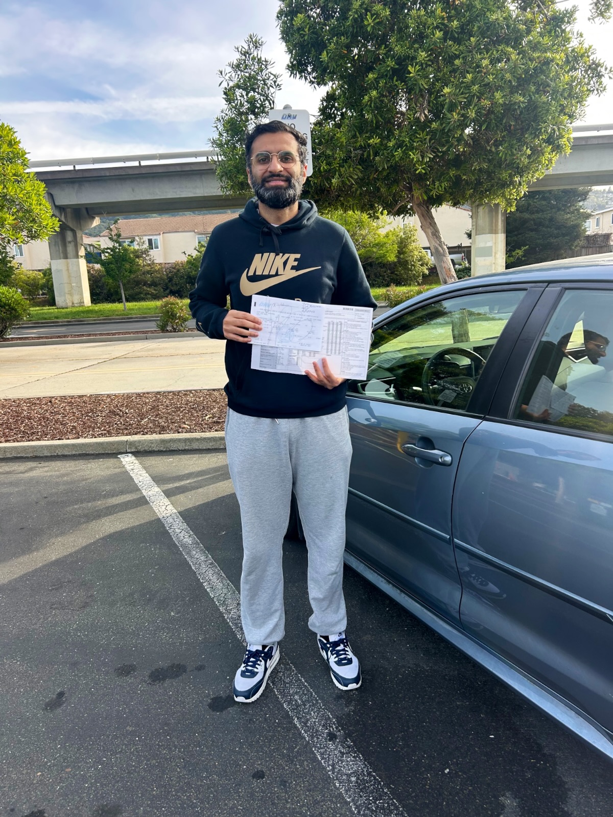 LLG Driving School student in a hoodie holding DMV drive test papers beside a blue sedan