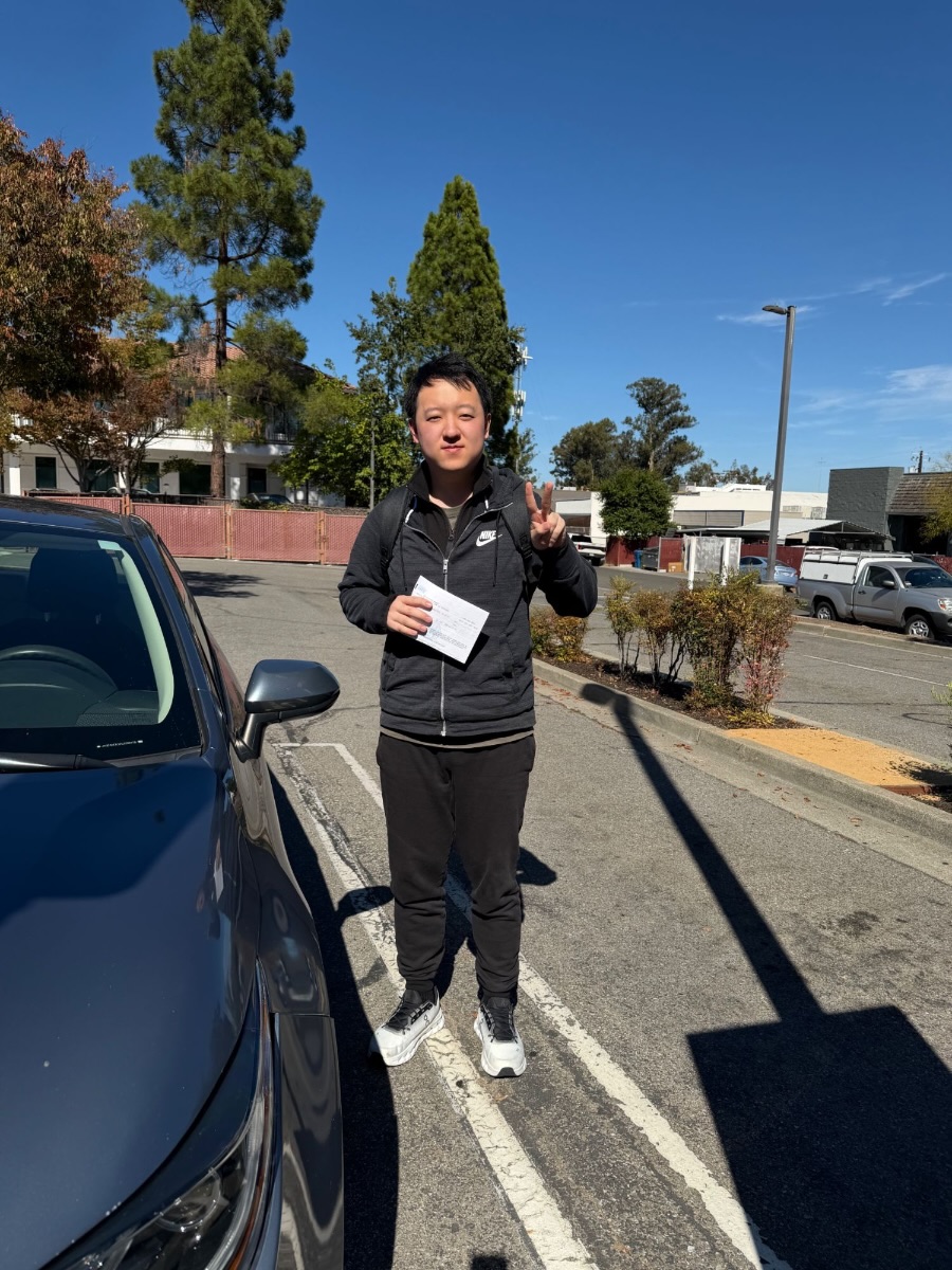 LLG Driving School student in a black jacket making a peace sign and holding a DMV receipt beside a car