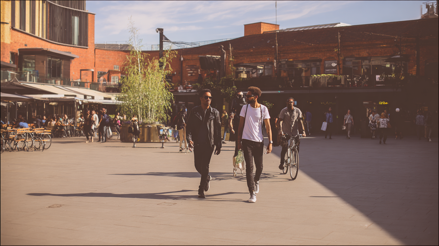 Two men walking and a man riding a bicycle in a sunny urban plaza with outdoor seating and brick buildings.