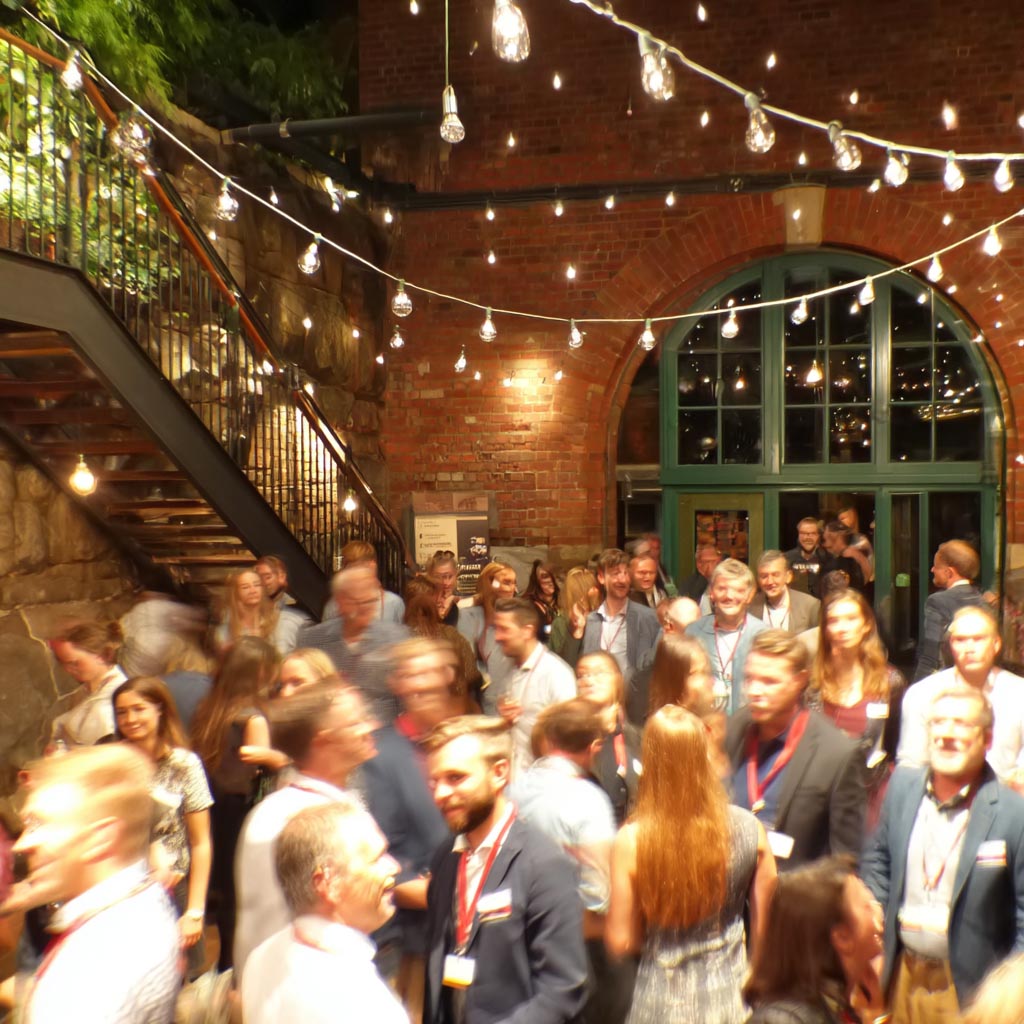 Crowd of people socializing in a warmly lit indoor space with string lights and brick walls.