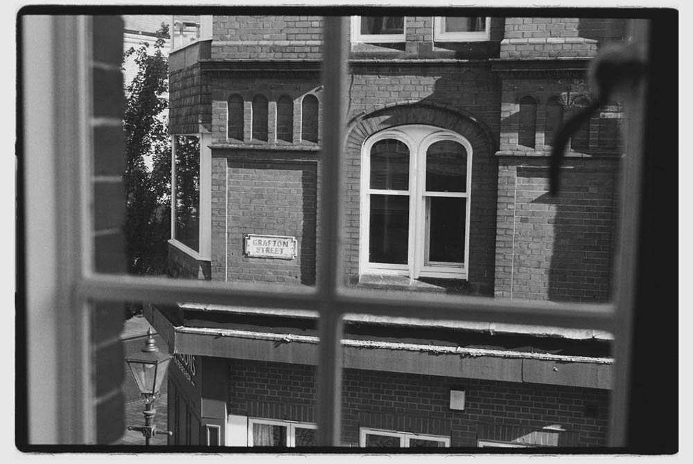 Black and white view through a window showing a brick building with arched windows and a Grafton Street sign.