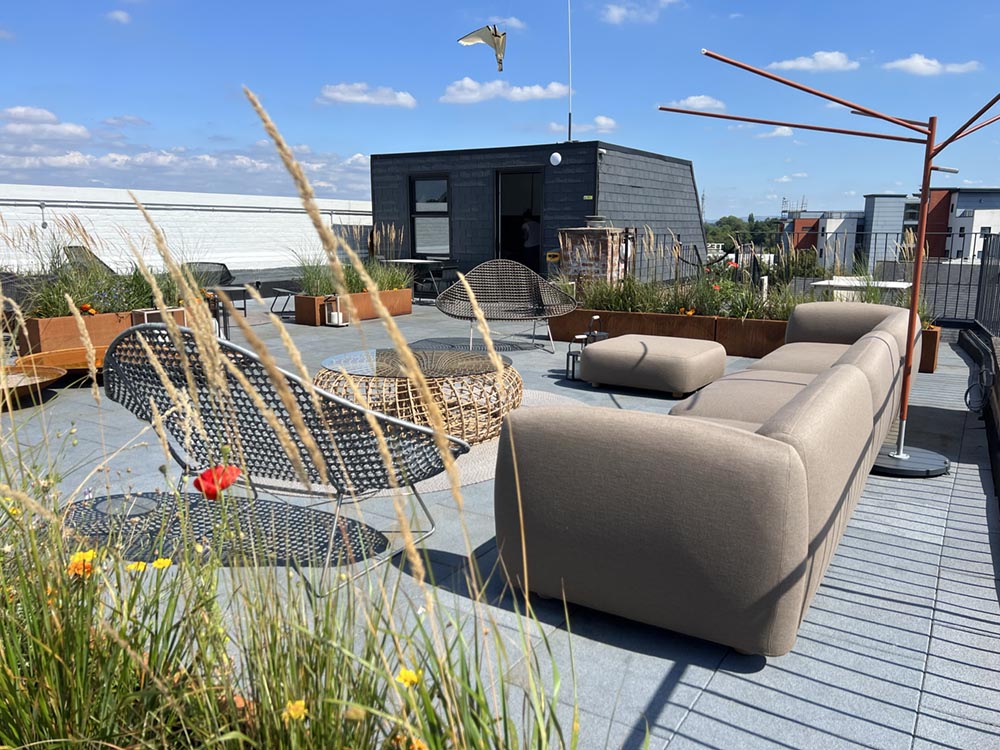 Modern rooftop patio with beige sectional sofa, metal wire chairs, round wicker coffee table, planters with ornamental grasses, and blue sky with scattered clouds.