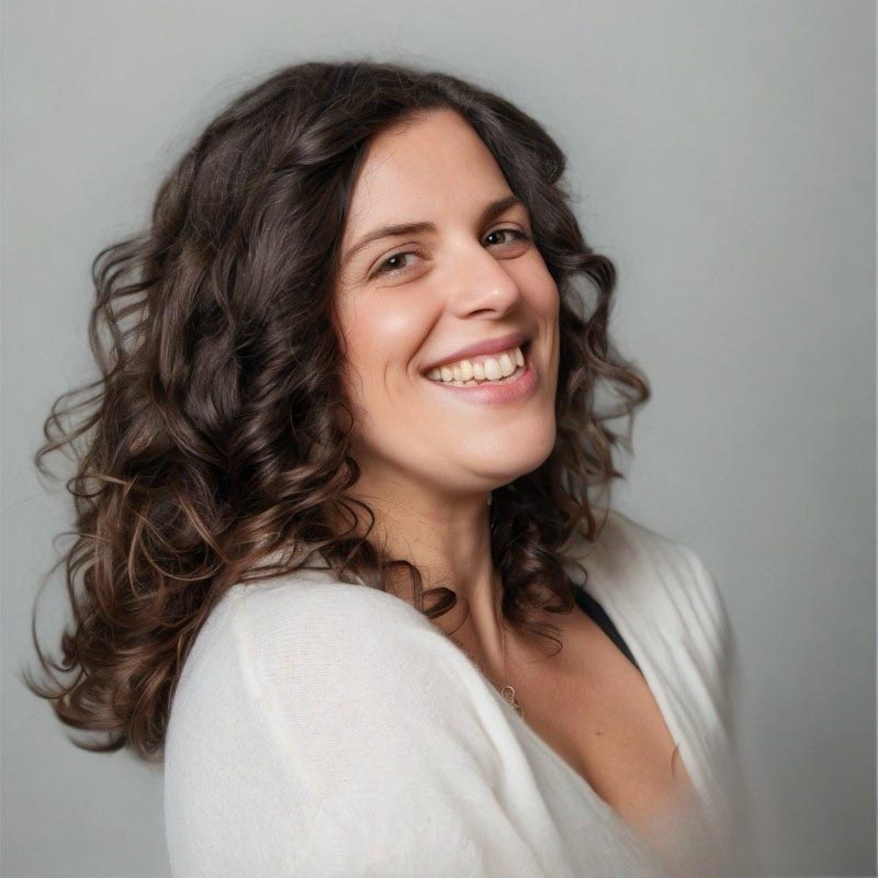 Smiling woman with curly dark hair wearing a white top against a light gray background.
