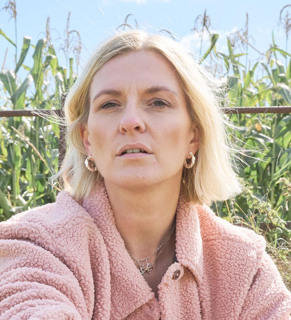 Blonde woman wearing a pink fluffy jacket and gold hoop earrings outdoors with green plants and blue sky in the background.