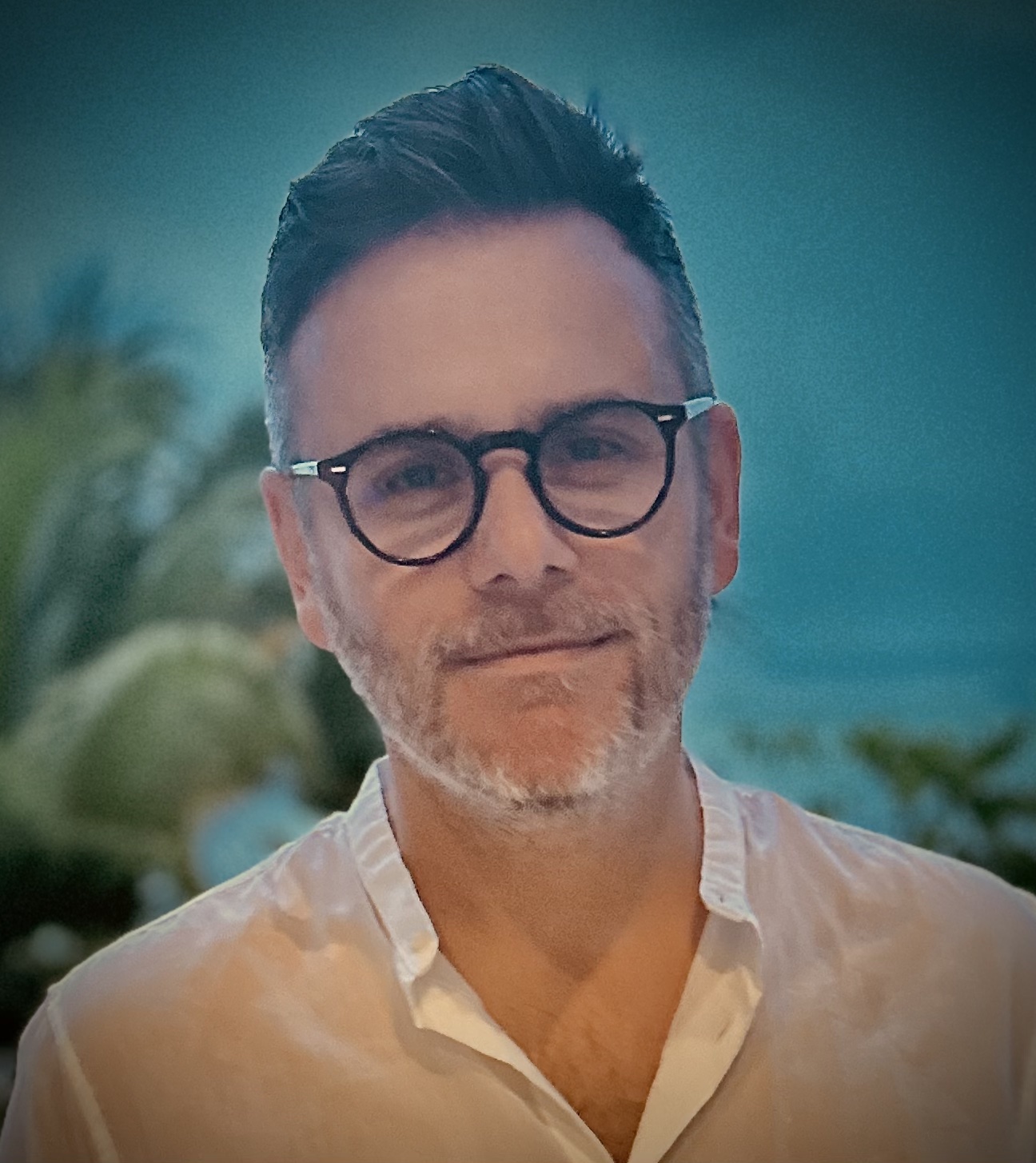 Man with short dark hair, beard, and round glasses wearing a white button-up shirt against a blurred outdoor background.