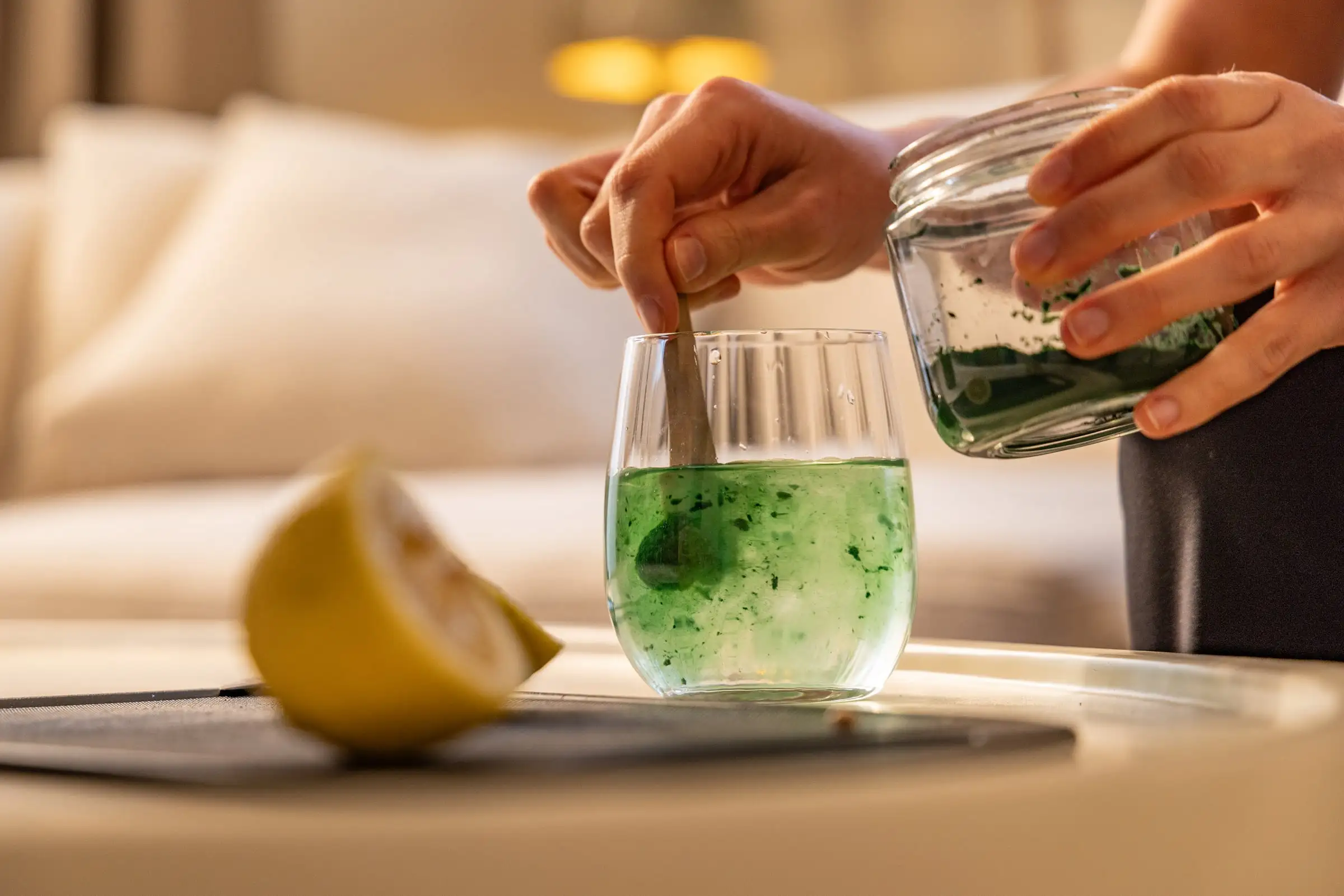 Ein Glas Wasser mit einer Zitrone neben dran und frischer Spirulina drin wird umgerührt. Die Rechte Hand rührt um während die Link hand das Spirulina Glas hält