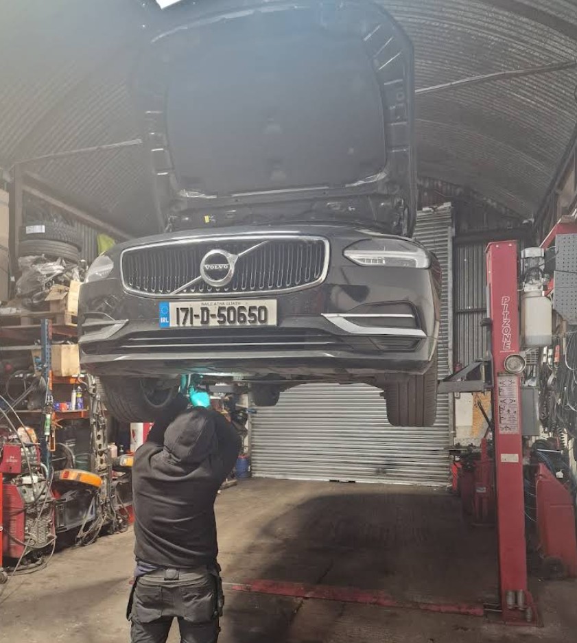 Mechanic working underneath a black Volvo car lifted on a red car lift inside a garage.
