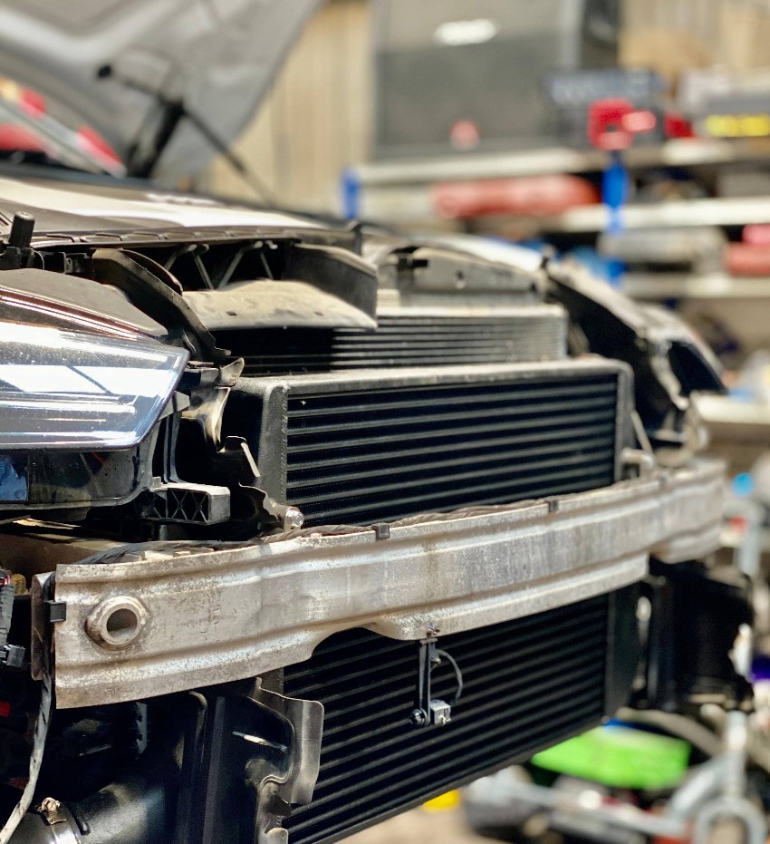 Close-up of a car front with the bumper removed, showing headlights, radiator, and metal support beam in an auto repair workshop.