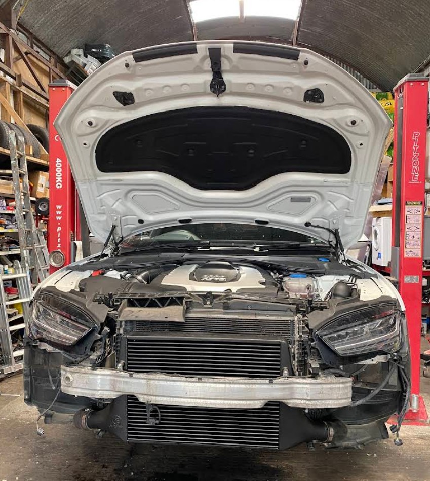 Front view of a white car with the hood open and the front bumper removed, showing the engine and radiator inside a workshop.