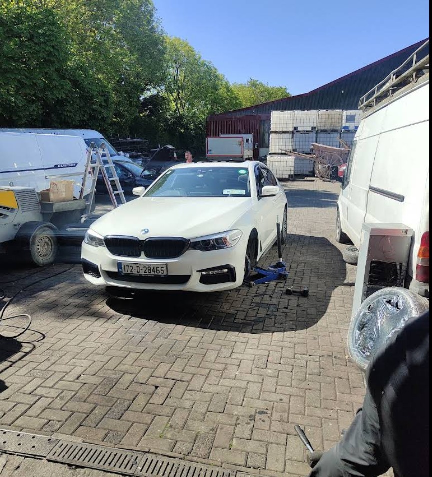 White BMW car raised on a blue jack in an outdoor workshop area with other vehicles, equipment, and stacked containers.