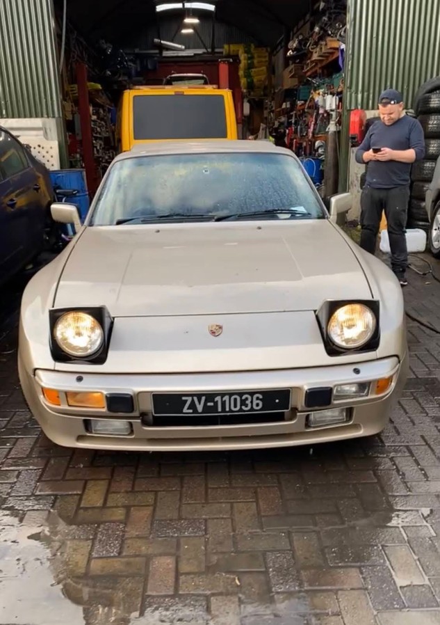 Front view of a classic silver Porsche 944 with raised headlights in a garage, with a person standing beside it looking at a phone.